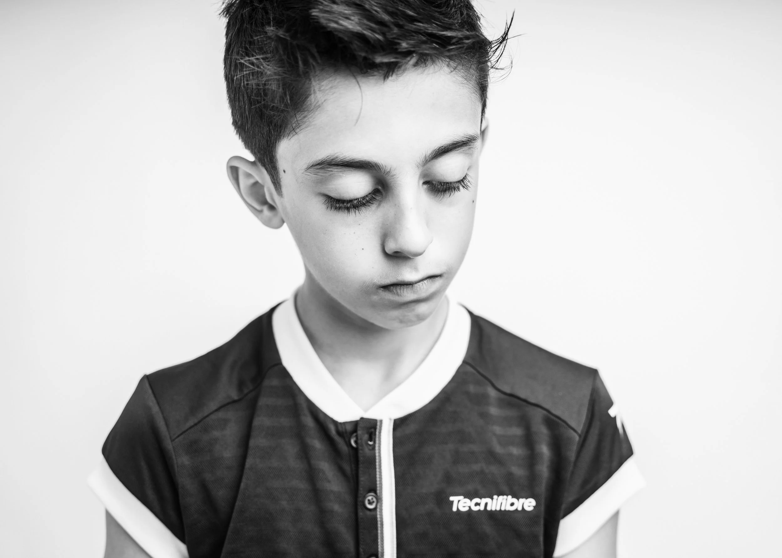 Black and white photo of a young man with short hair and closed eyes, wearing a sports jacket with a 'Tecnifibre' logo, against a plain background.