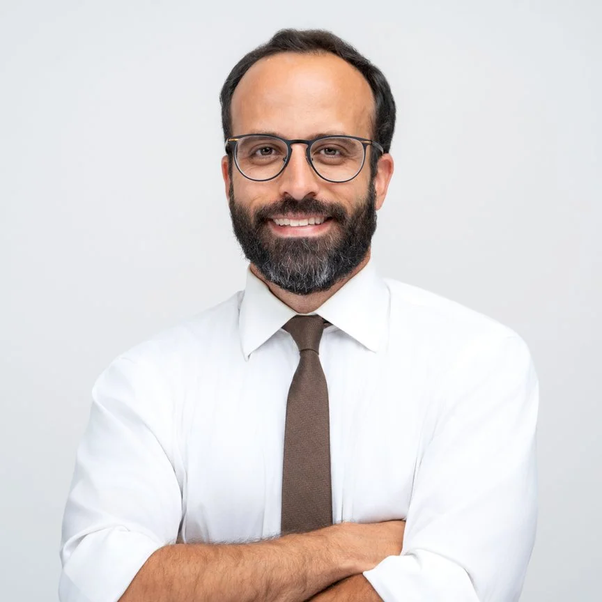 A smiling man wearing glasses, a white dress shirt, and a brown tie, standing against a plain light gray background.