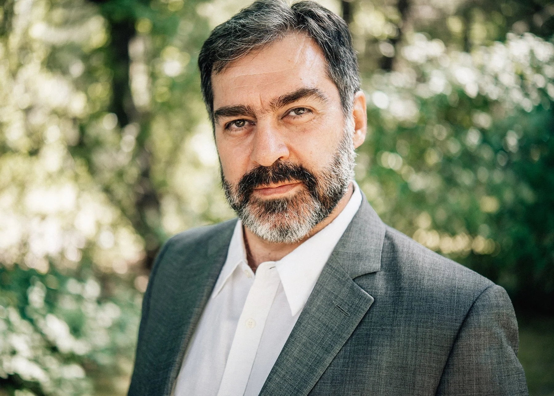 A middle-aged man with dark hair and a beard, wearing a gray suit jacket and white shirt, standing outdoors in a natural setting with blurred green foliage in the background.