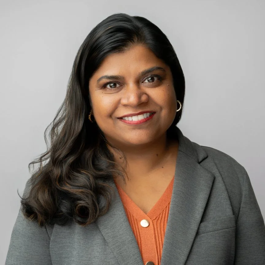 A woman with long, dark, wavy hair, wearing a gray blazer over an orange sweater with a button, smiling, against a plain gray background.