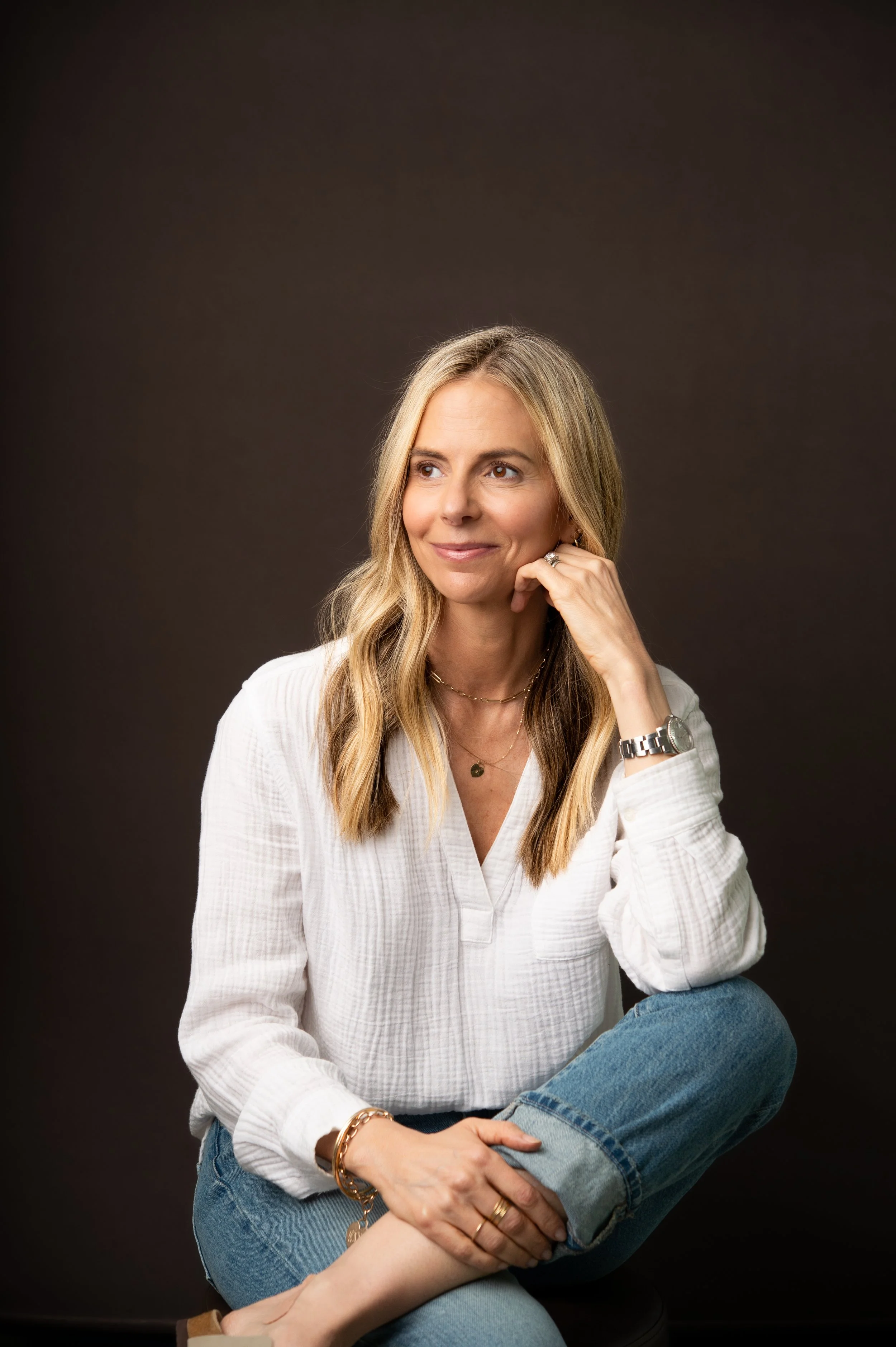 A woman with blonde hair sitting with her leg crossed on her knee, wearing a white blouse and blue jeans, smiling and looking to her left, against a dark background.