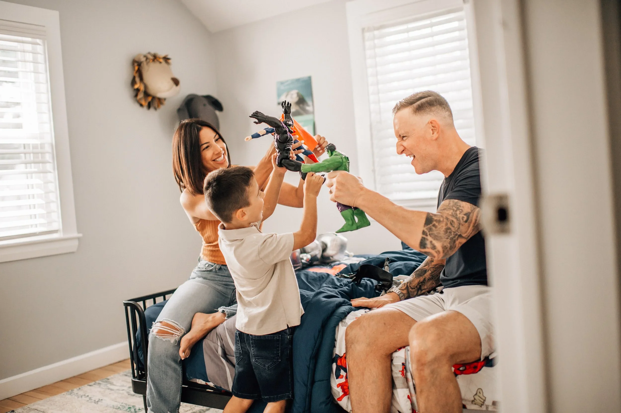 A family of four having fun on a bed in a bright bedroom, playing with action figures. The mother, father, and two children are smiling and engaging in playful activity with their toys.