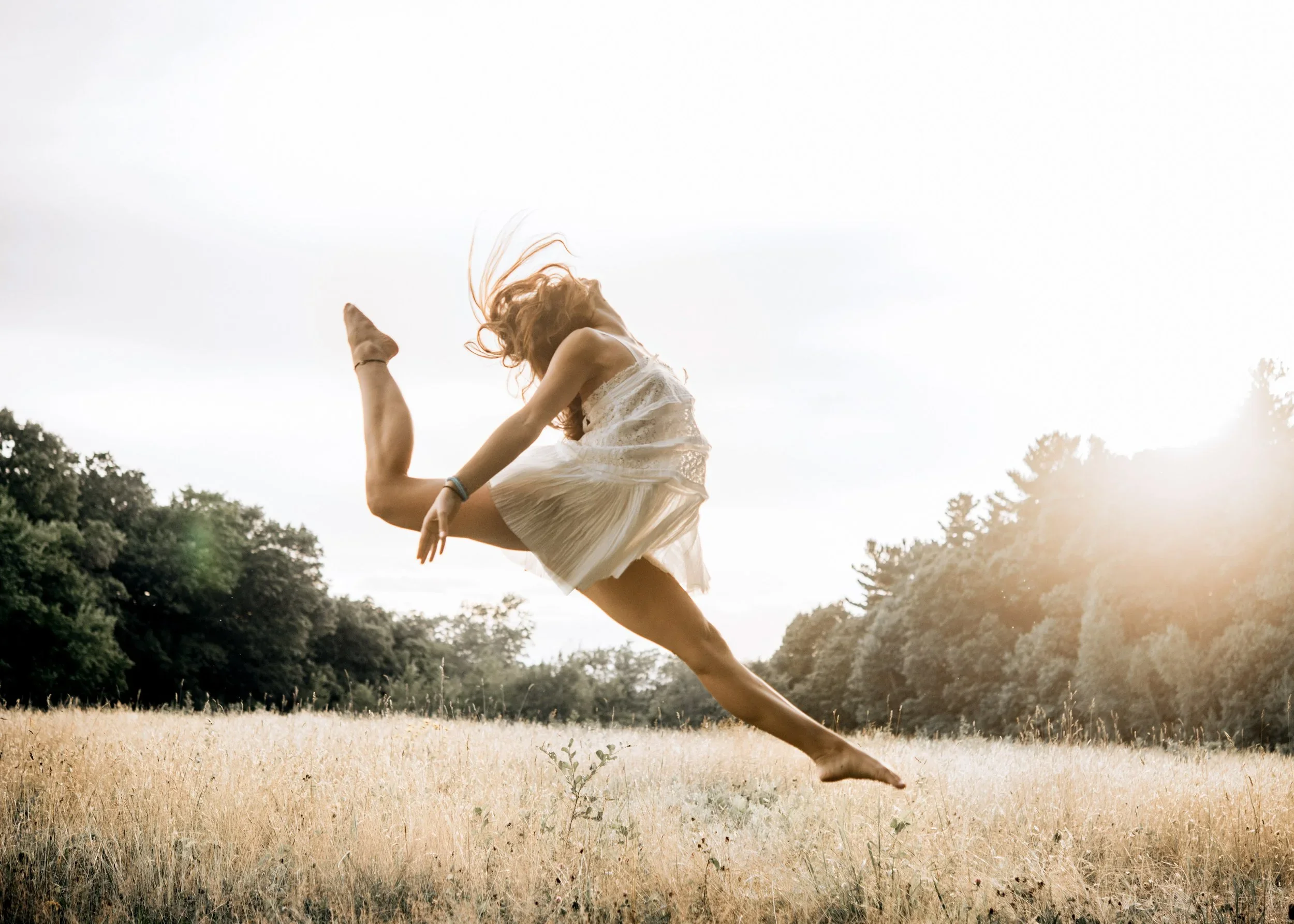 A woman jumping and stretching in a field with trees in the background at sunset.
