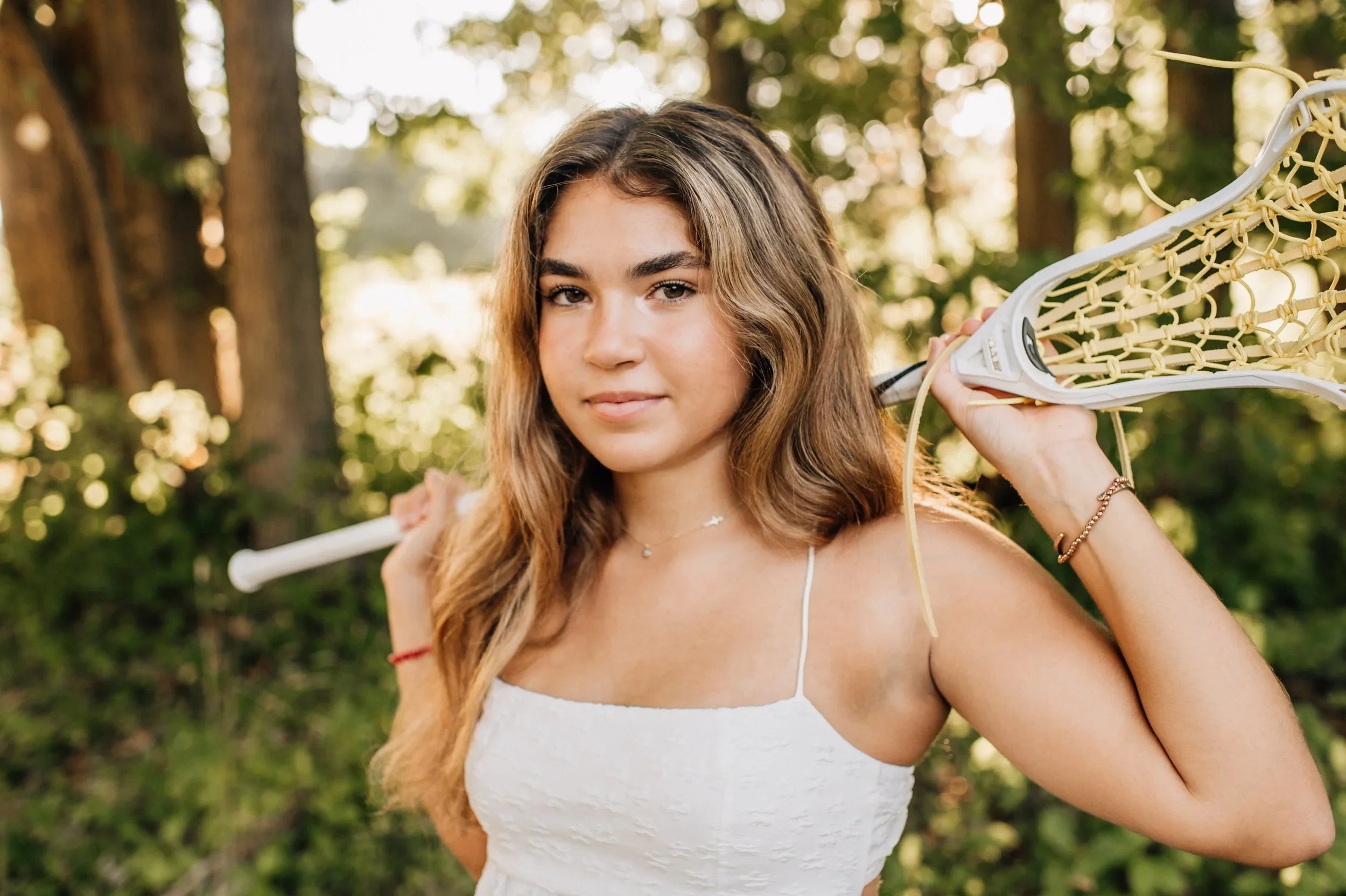 A young woman with long, wavy hair stands outdoors in a wooded area, holding a lacrosse stick over her shoulder. She is wearing a white sleeveless top and has jewelry including a bracelet and a necklace, with a subtle smile and natural background of trees and sunlight.