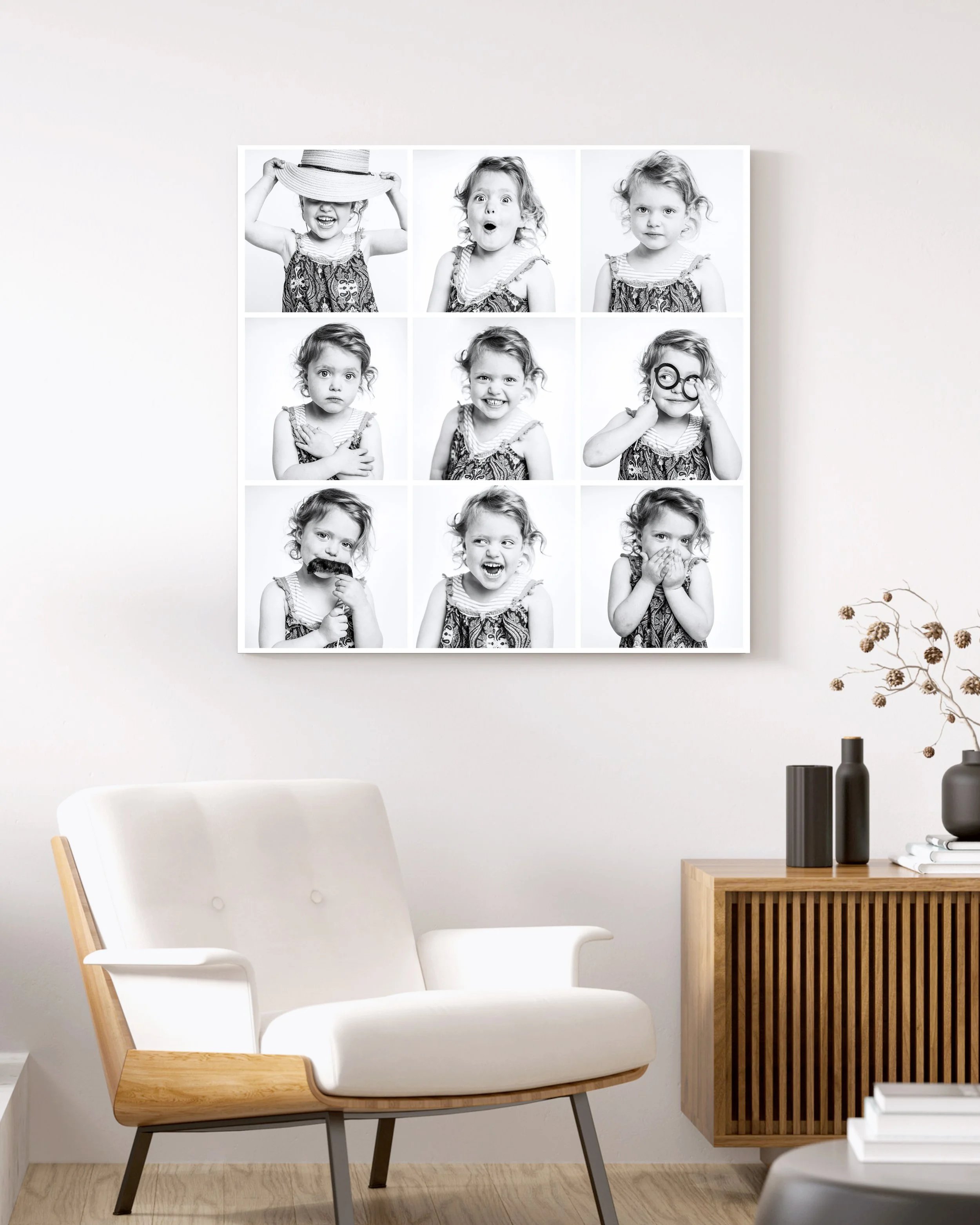 A white wall with a black and white photo collage of a young girl making different facial expressions. A modern white chair with wooden arms and gray legs is in the foreground, and a wooden side table with black and white vases and dried flowers is on the right.