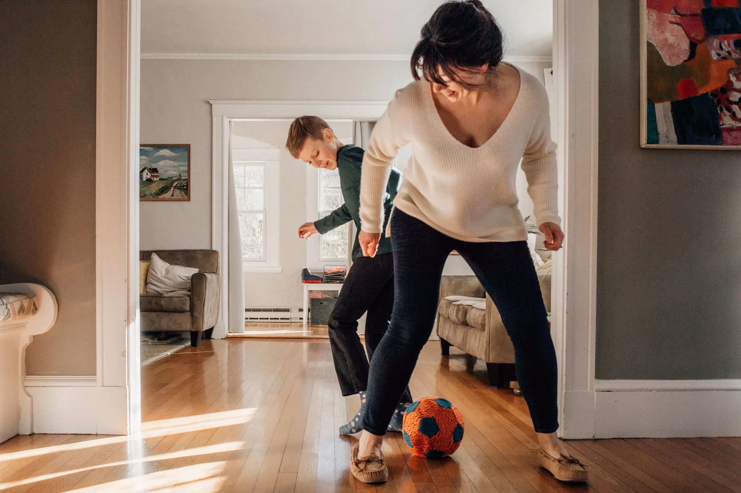A woman and a young boy playing indoor soccer with an orange and black ball in a living room.
