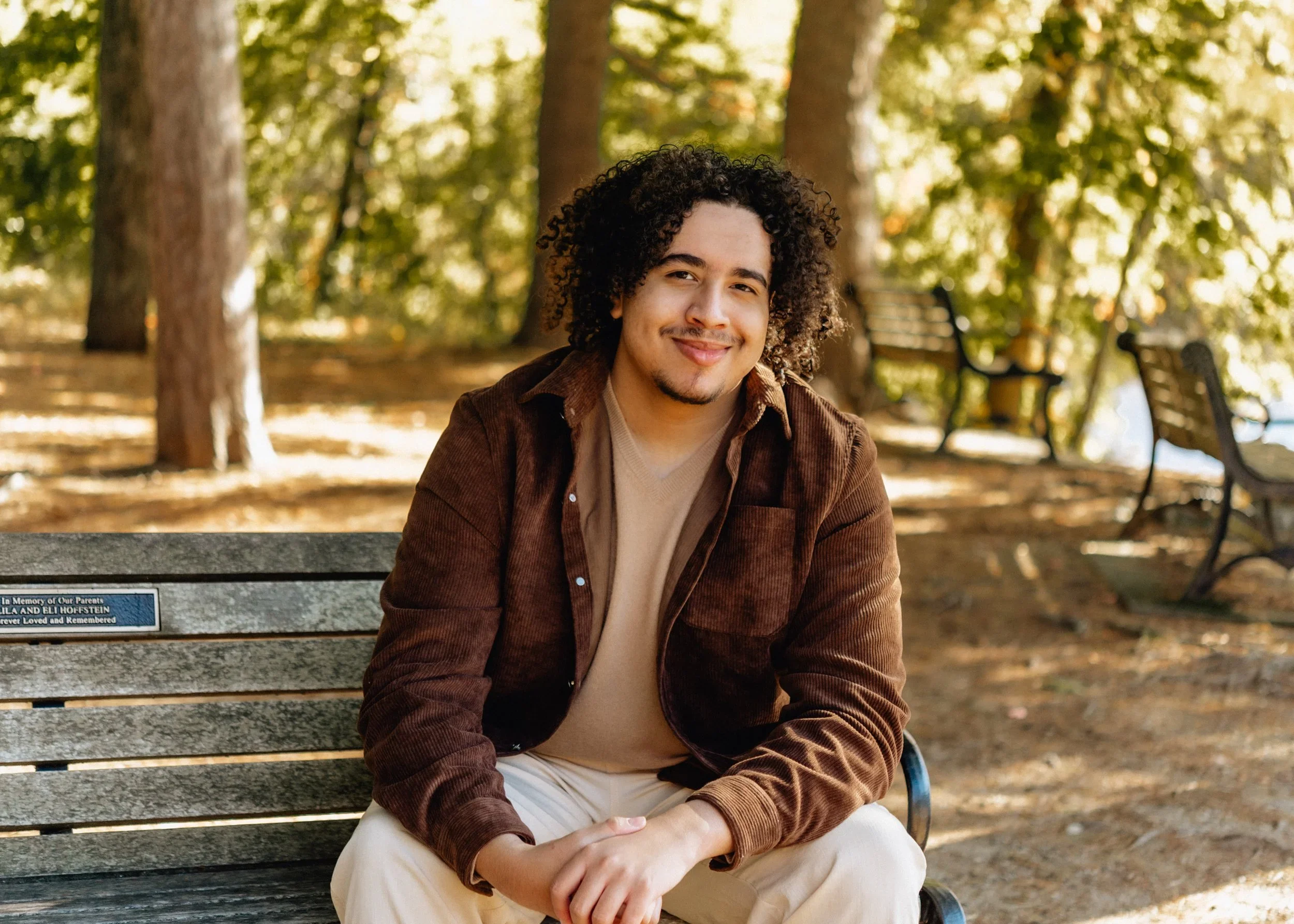 Young man with dark curly hair and a goatee, smiling, sitting on a park bench with trees and benches in the background during autumn.