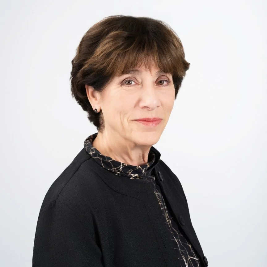 Portrait of a middle-aged woman with short brown hair, wearing a black blazer and patterned shirt, smiling softly against a white background.
