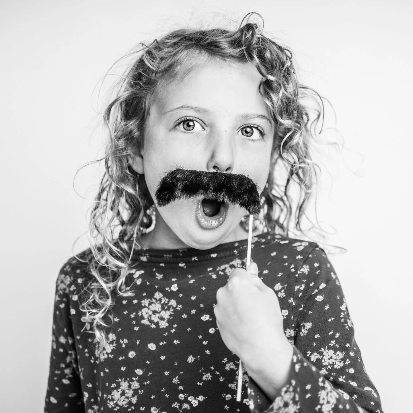 A young girl with curly hair holds a fake mustache on a stick in front of her face, with her mouth open in surprise or excitement.