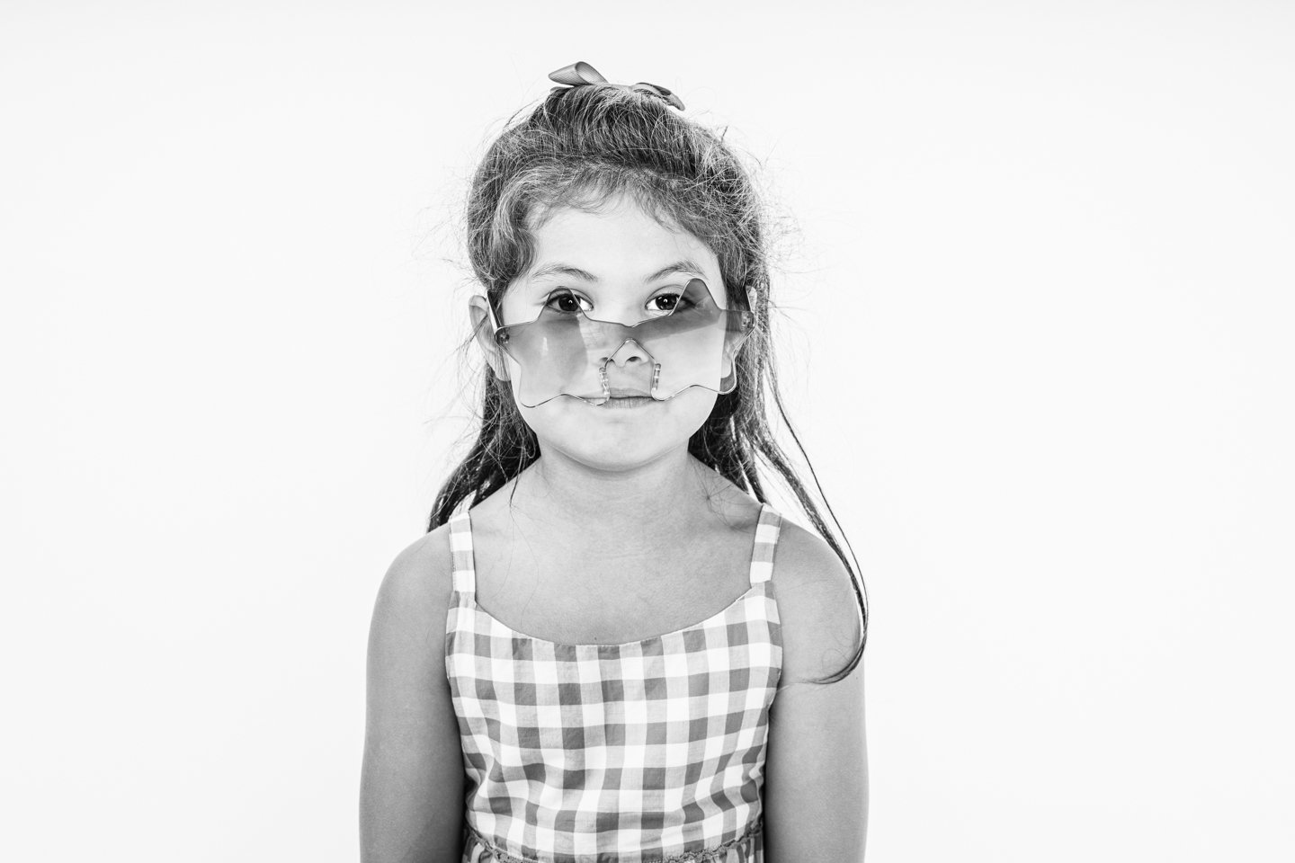 A young girl wearing star-shaped sunglasses and a gingham dress, smiling with her hair in a ponytail with a bow, against a plain white background.