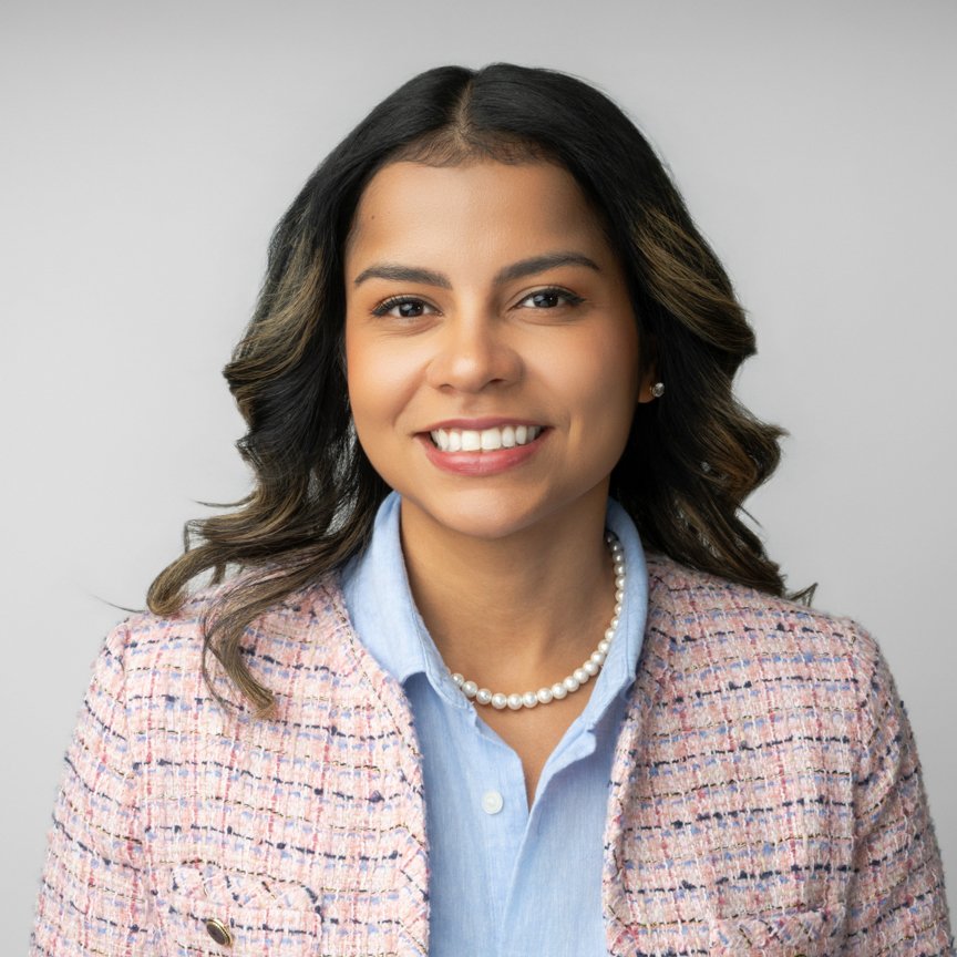 A woman with shoulder-length dark hair, smiling, dressed in a light blue collared shirt and a pastel-colored tweed blazer, wearing pearl earrings and a pearl necklace, against a plain light gray background.