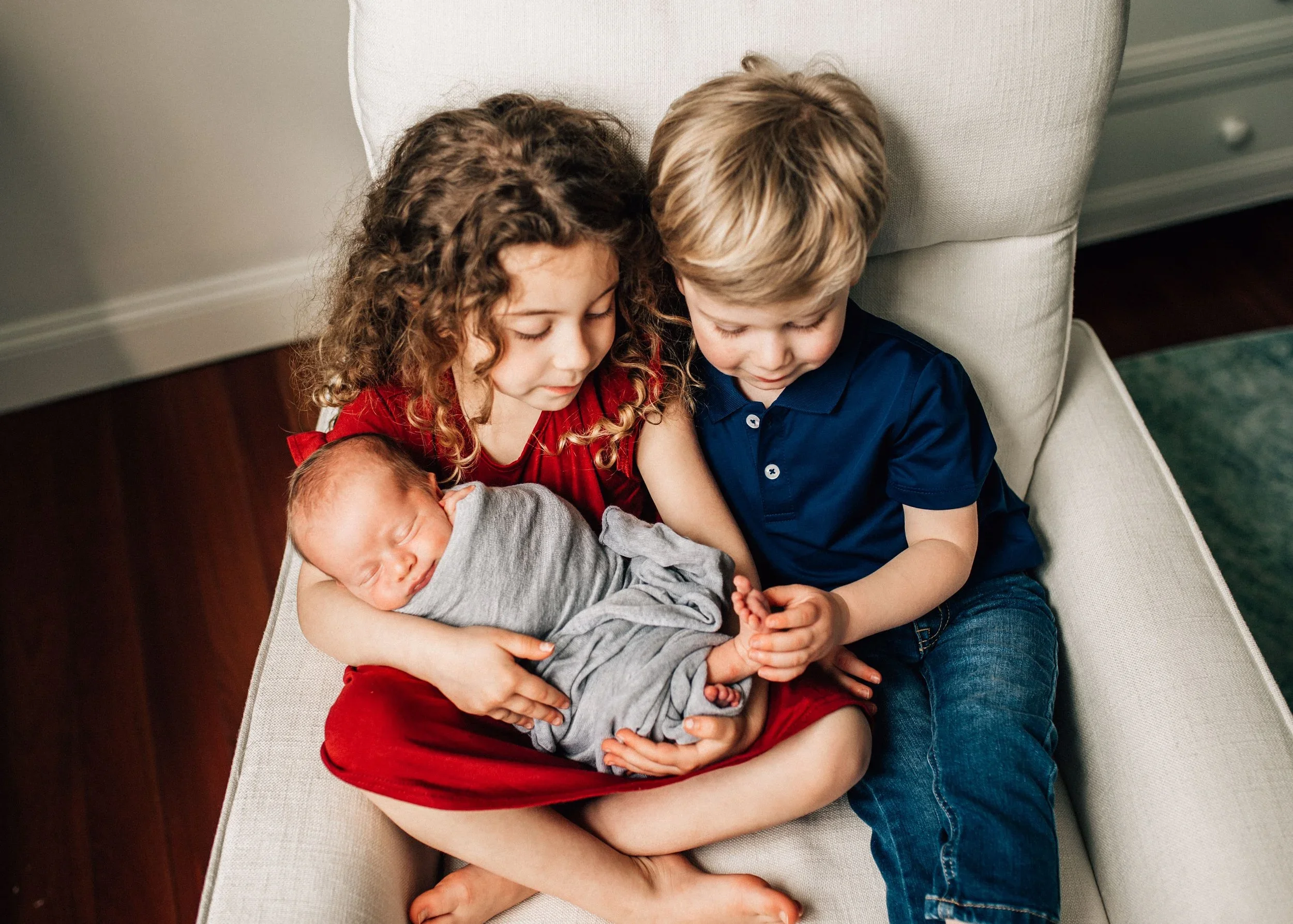 A young girl, a young boy, and a newborn baby are sitting on a light-colored sofa. They are closely gathered, with the children looking down softly at the baby, who is wrapped in a gray blanket and wearing a gray outfit. The girl has curly hair and is wearing a red dress, while the boy has straight blond hair and is dressed in a navy blue polo shirt and jeans.
