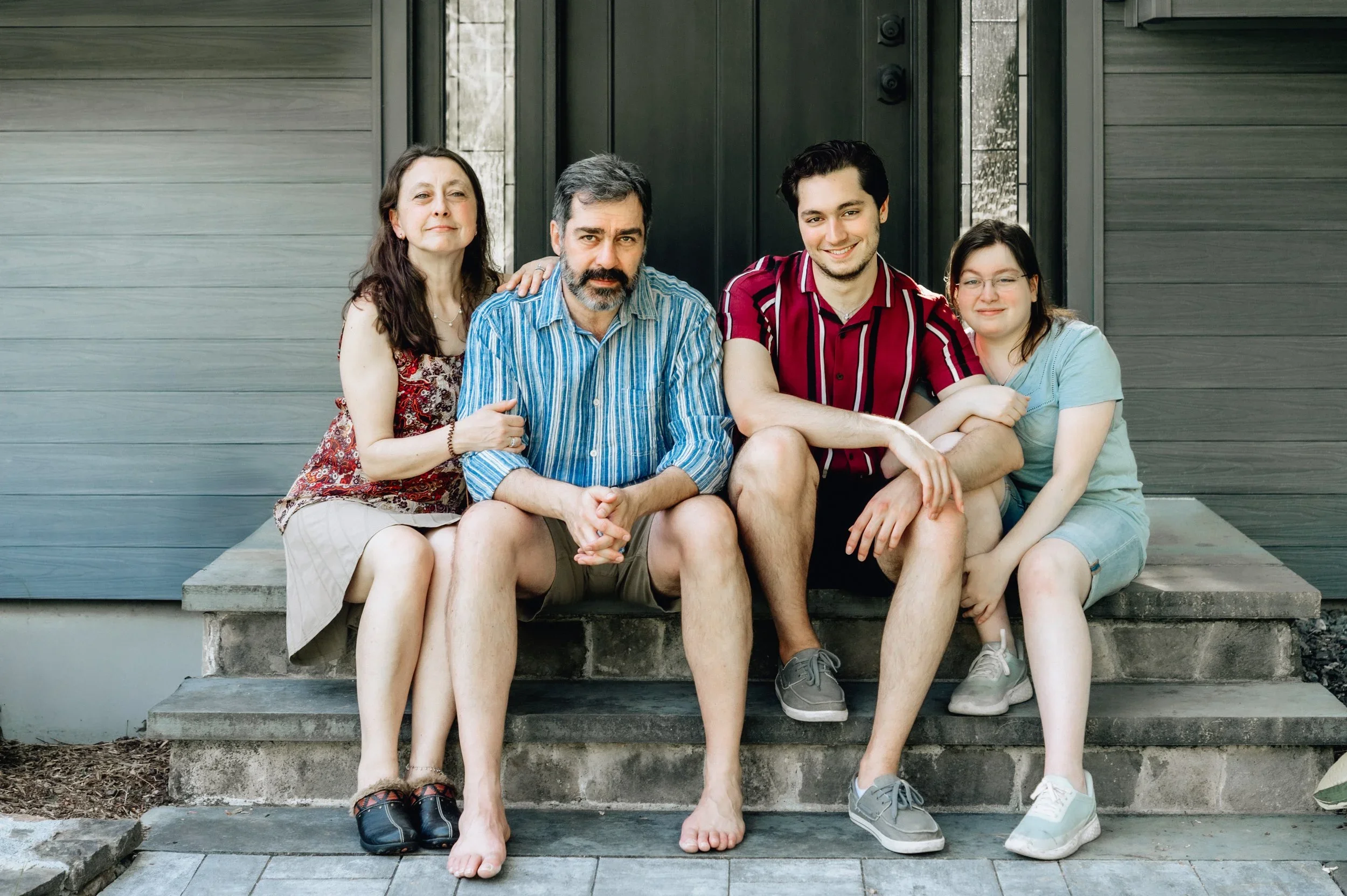 A multi-generational family of four sitting on front steps outside of a house, smiling and relaxed.