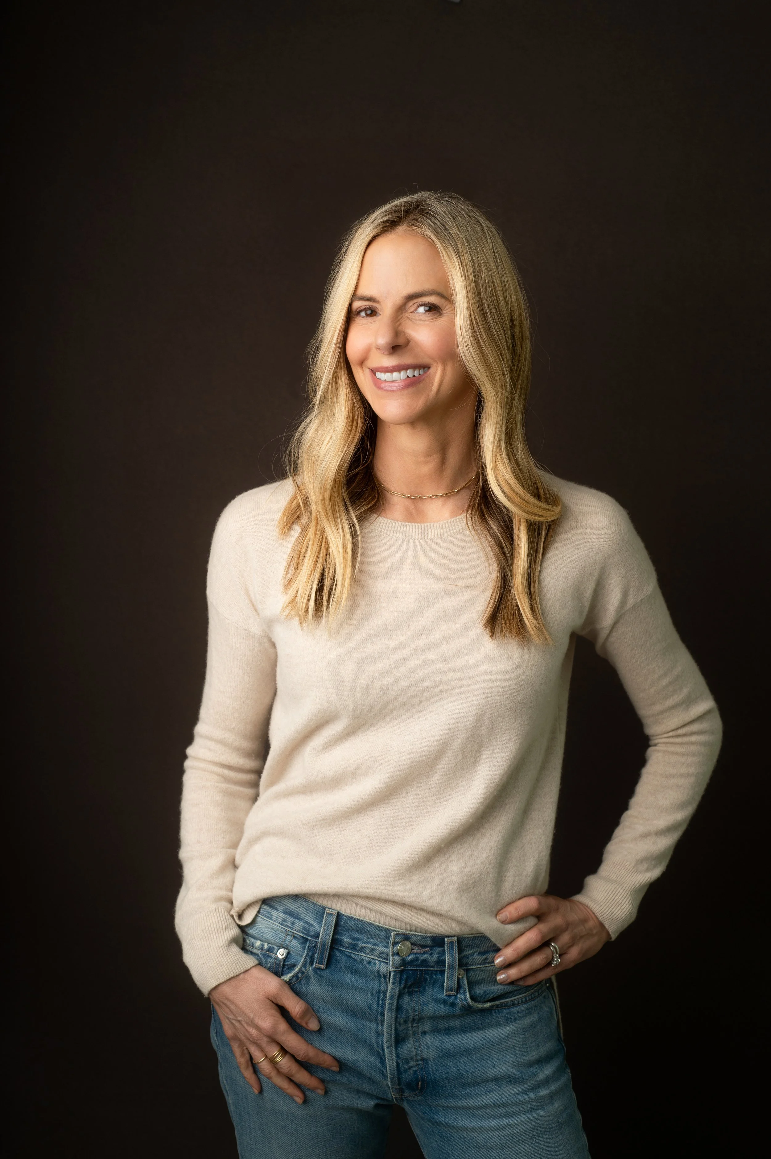 A woman with blonde hair, wearing a beige sweater and blue jeans, standing against a dark background, smiling and looking at the camera.