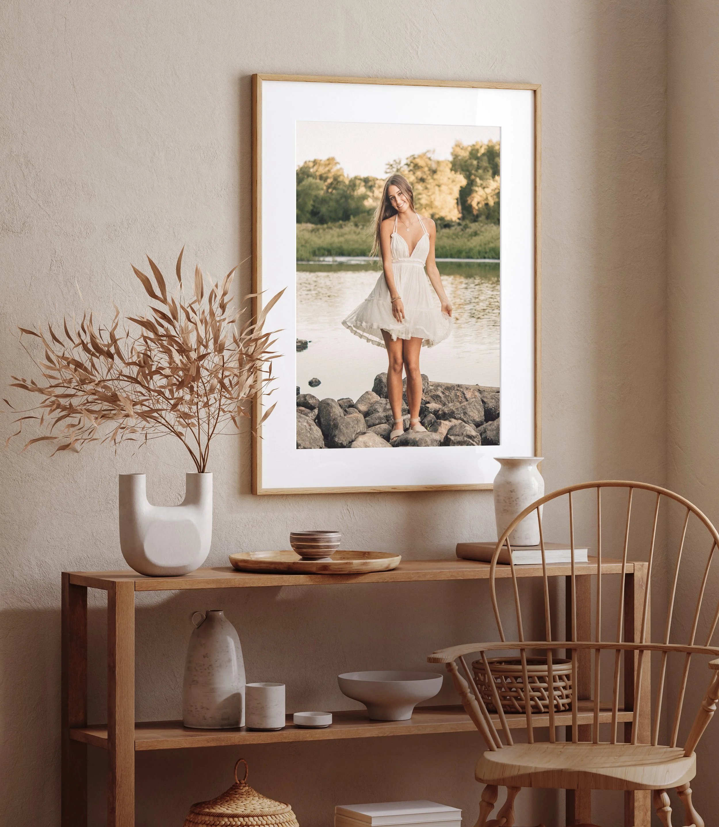 Decorative interior with a framed photo of a woman by a river on a beige wall, wooden table with white ceramic vases of dried plants, bowls, and books, a rattan chair and woven basket.