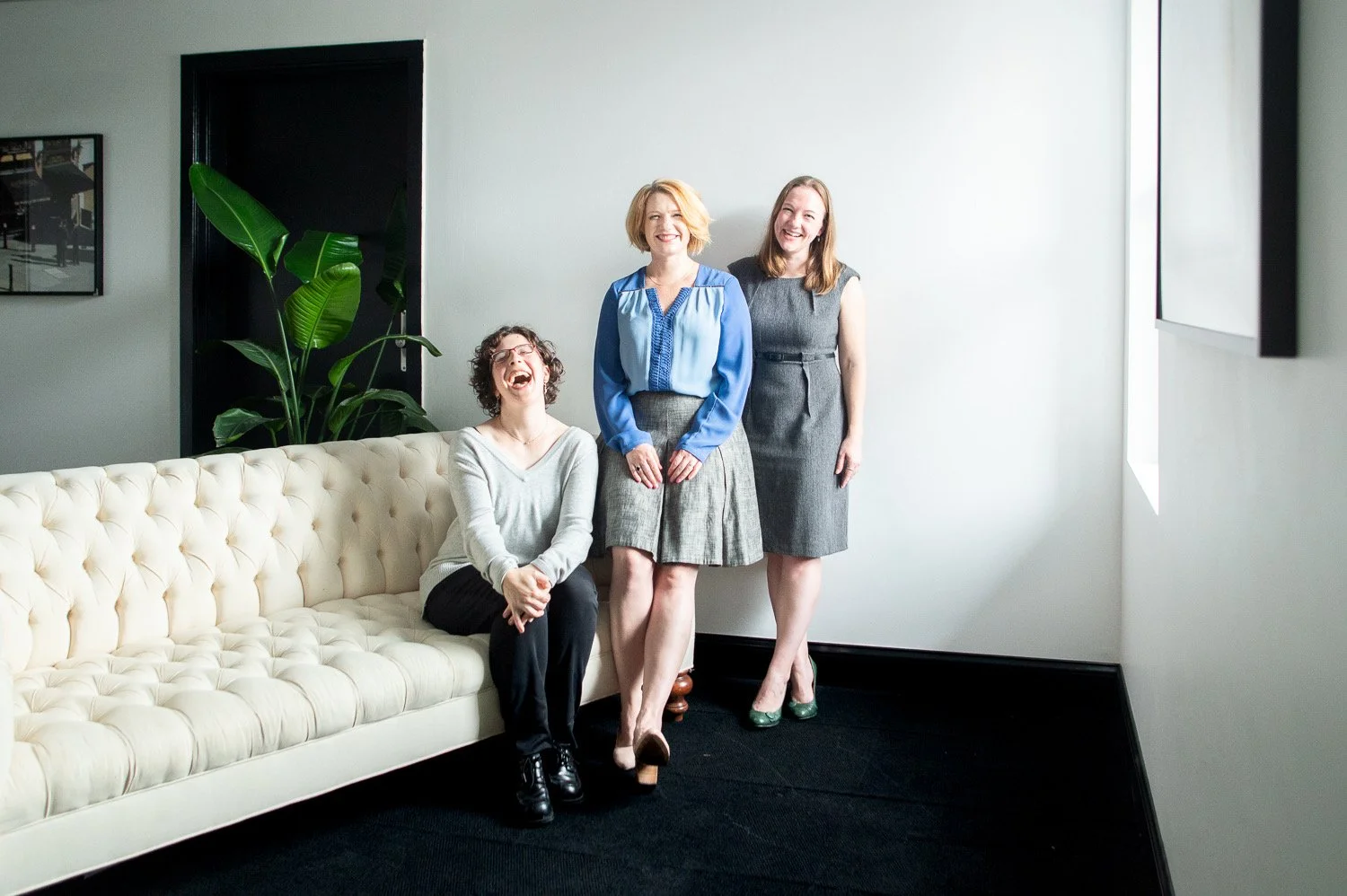 Three women laughing and smiling in a modern room, standing near a white tufted sofa, with a tall green plant and framed art on the wall.