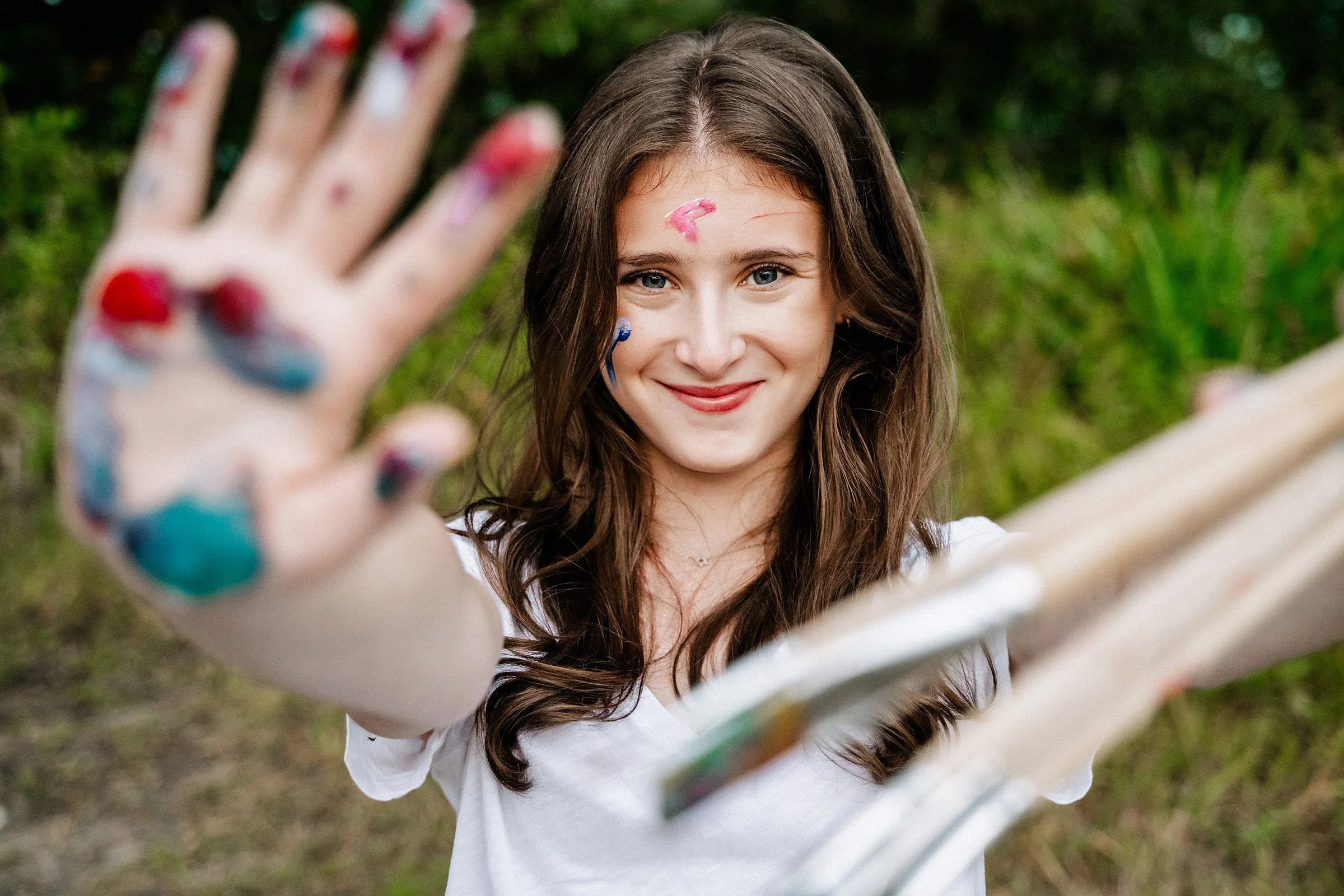 Young girl with painted face and hands reaching out towards the camera, outdoors with green foliage in the background.