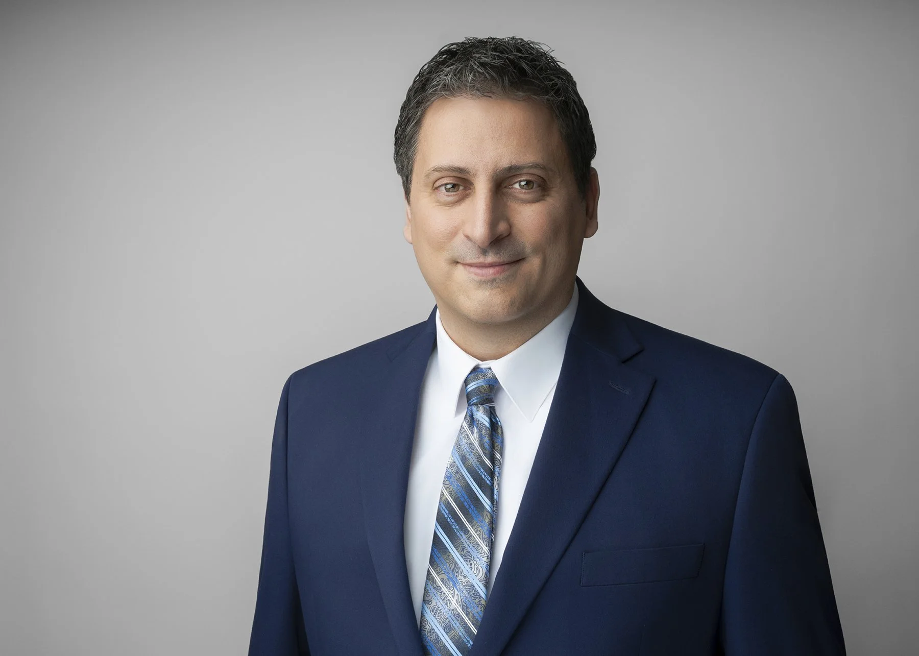 A professional man in a navy suit, white shirt, and patterned tie smiling against a gray background.