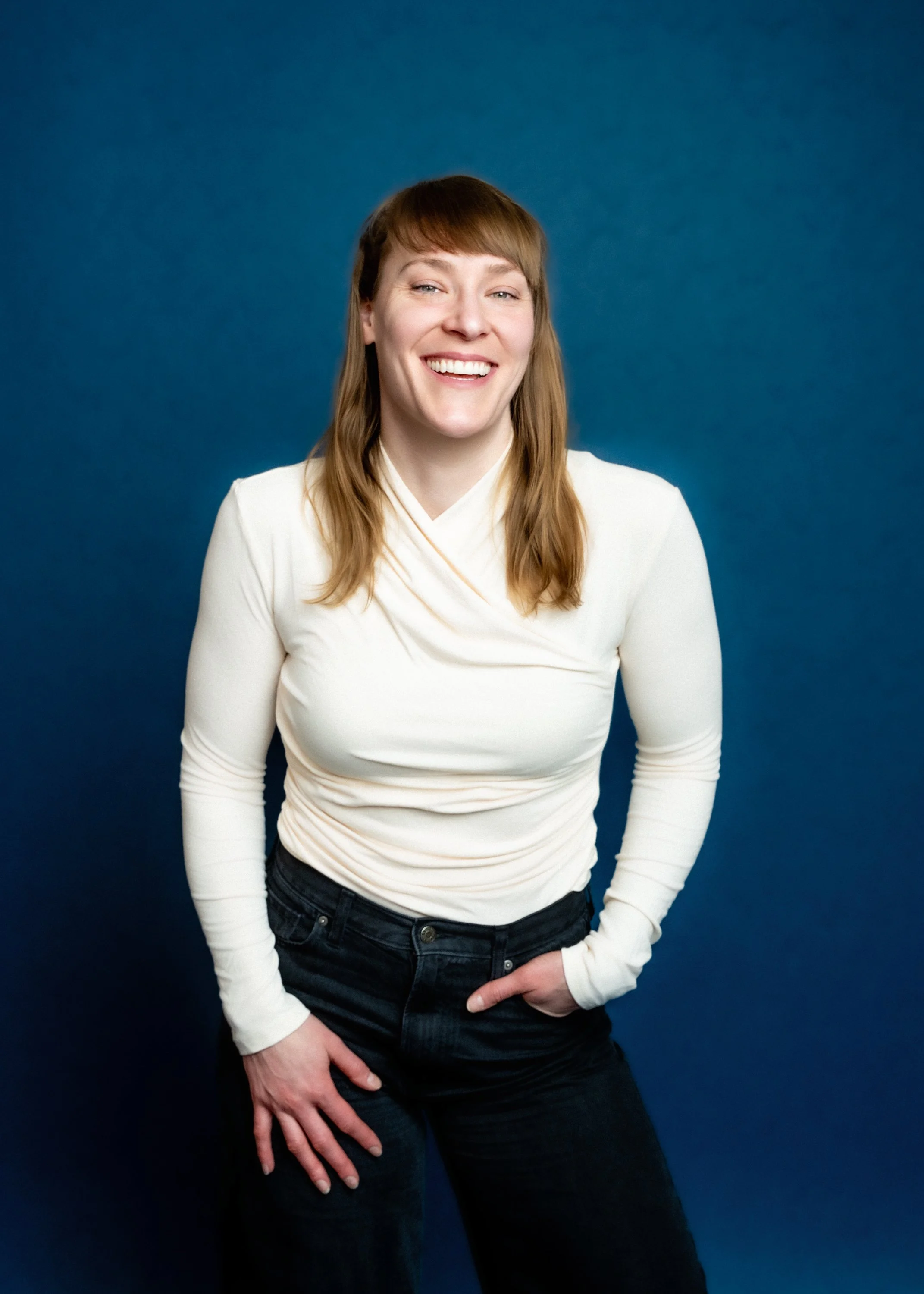 A woman with long, brown hair wearing a white, long-sleeved top and dark jeans, standing against a solid blue background, smiling with her mouth open.
