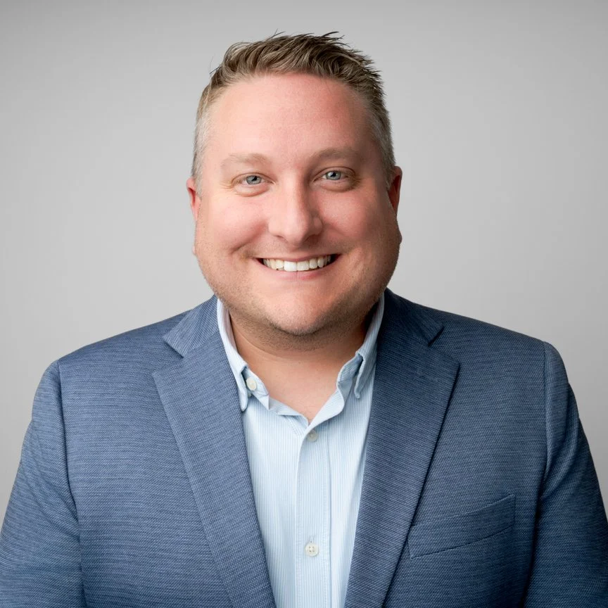 A smiling man in a gray blazer and light blue button-up shirt facing the camera against a plain gray background.