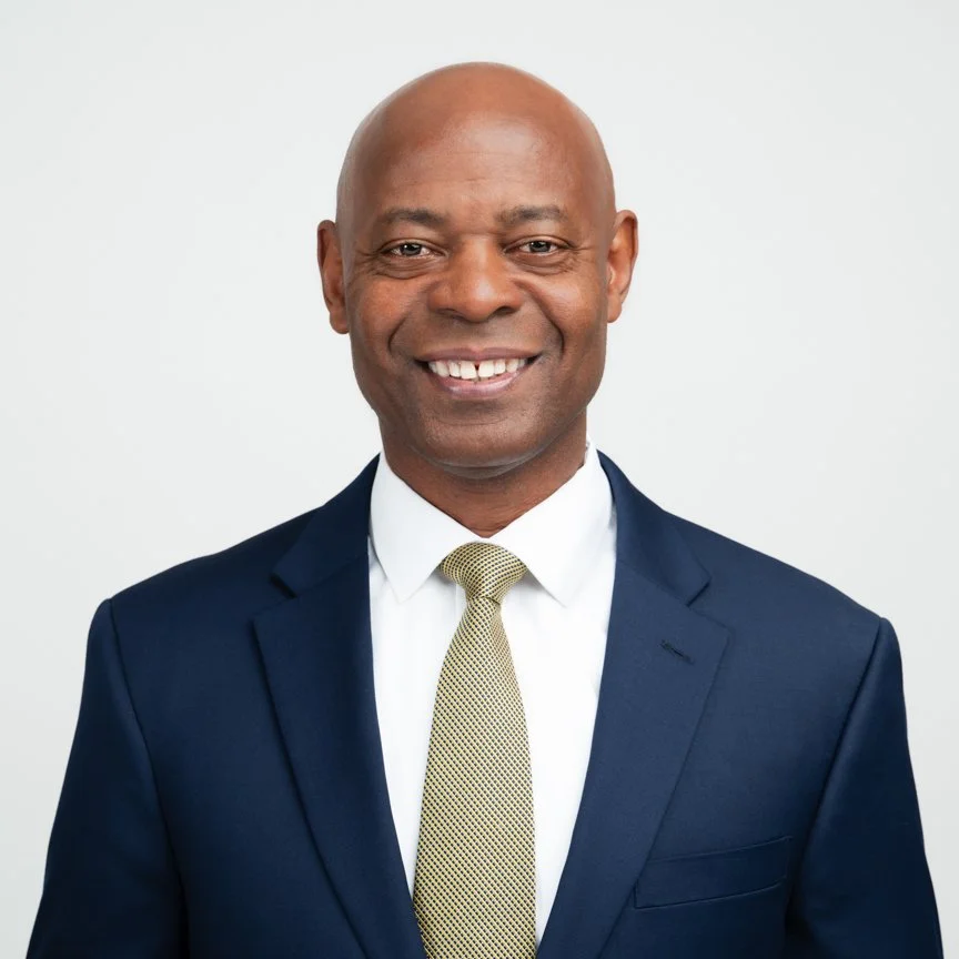 A professional man wearing a dark suit, white shirt, and yellow tie, smiling against a plain light background.