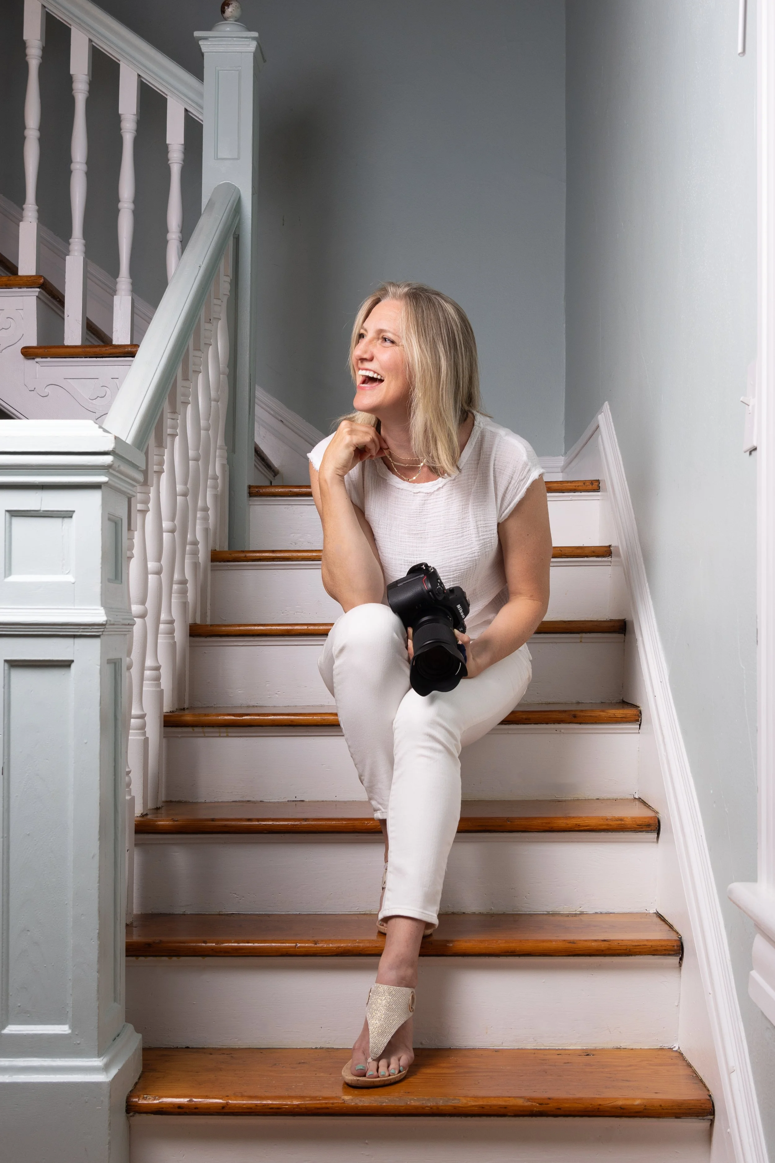 A woman with blonde hair sitting on wooden stairs, holding a camera in her lap, wearing a white top and white pants, smiling and looking to her left.