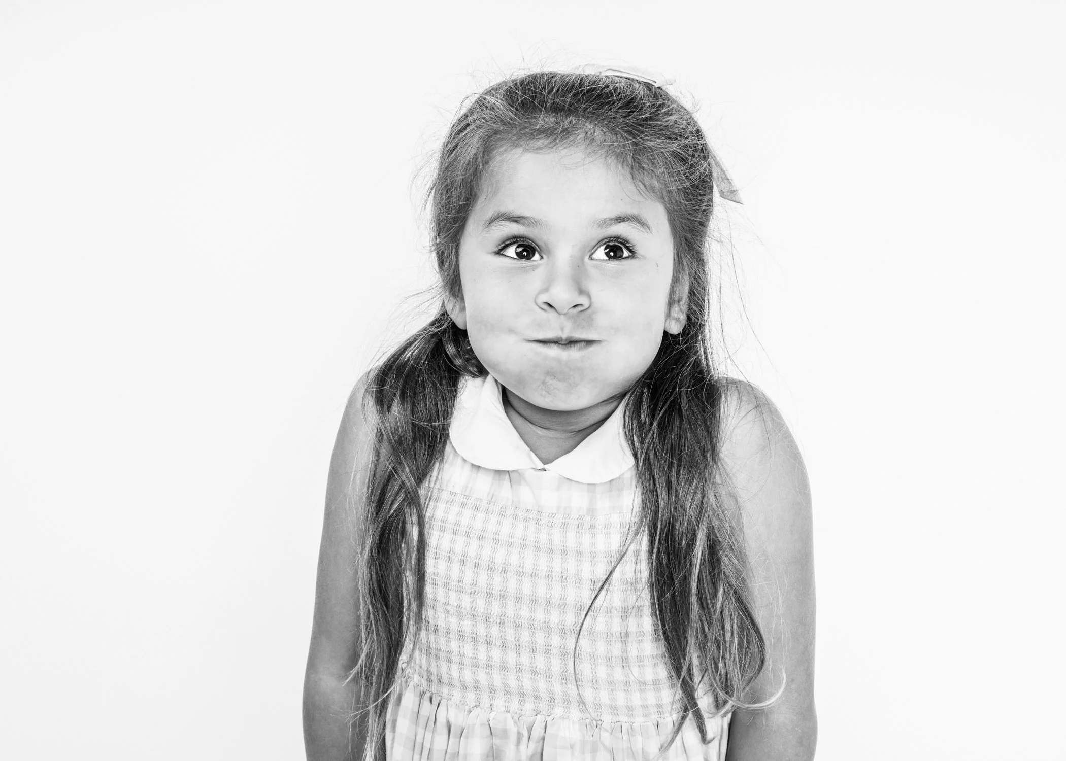 Black and white photo of a young girl with long hair, wearing a striped sleeveless top, making a playful facial expression with puffed cheeks and wide eyes.