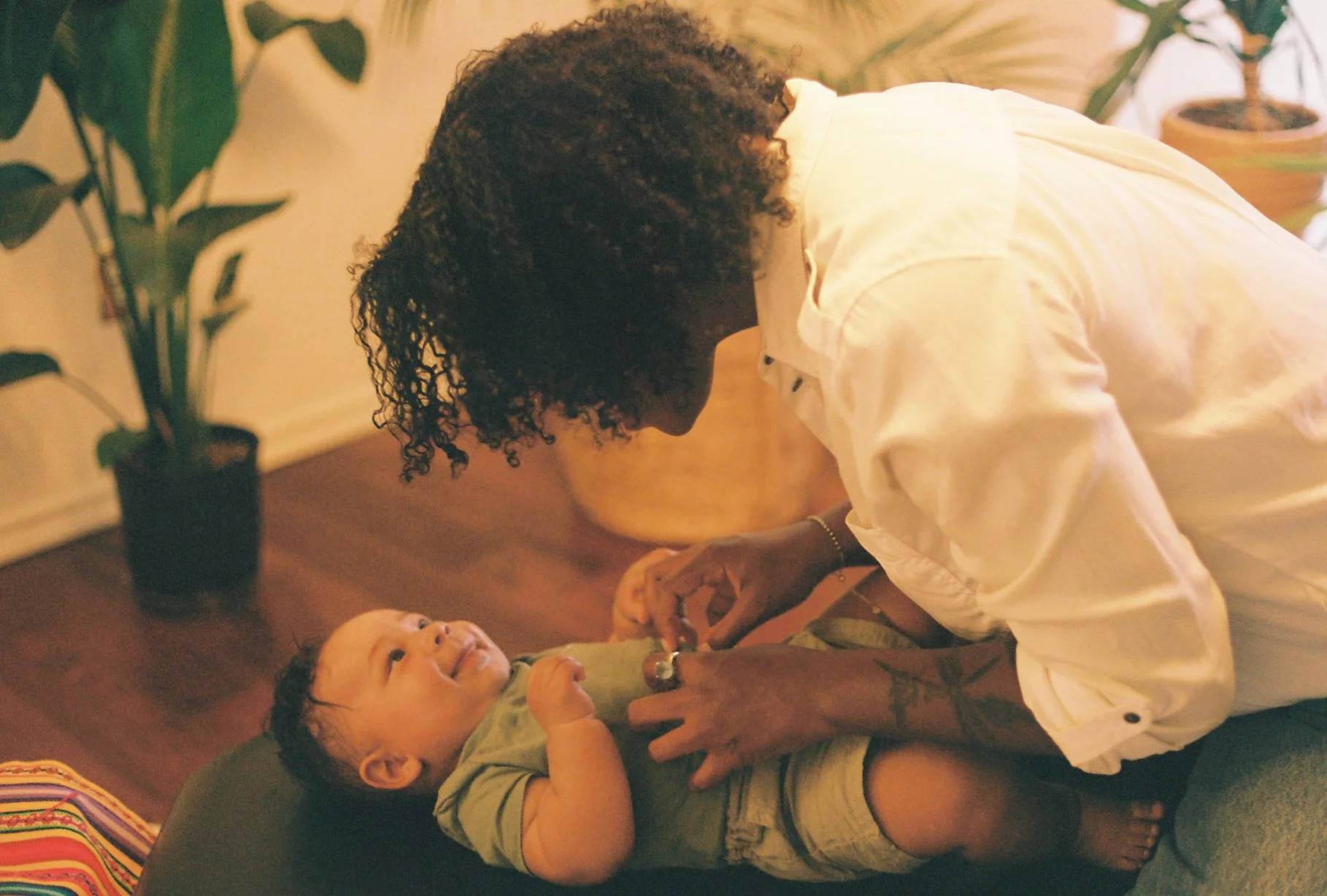 A woman with curly hair leaning over a baby who is lying on a soft cushion, holding the baby's hands in a cozy indoor setting with plants in the background.