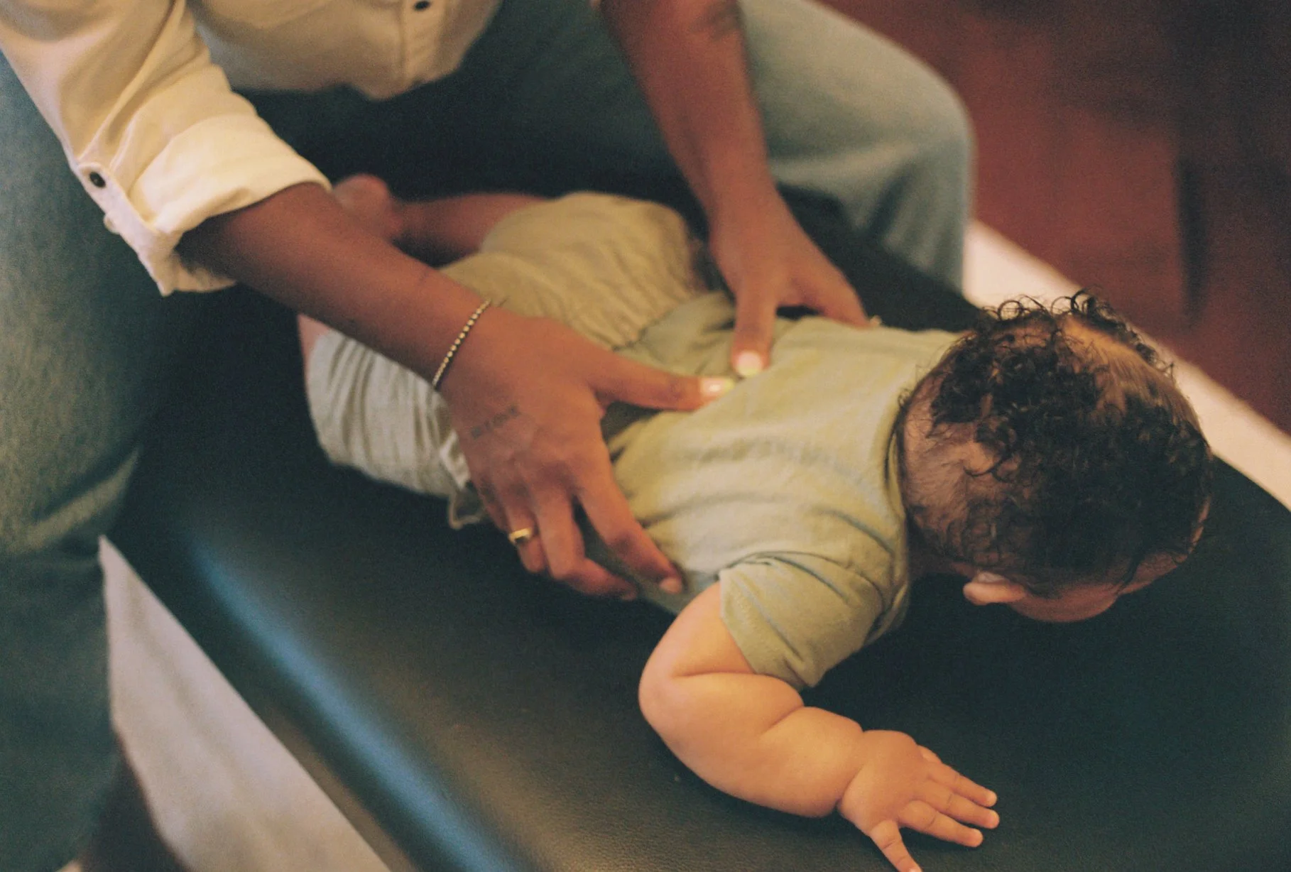 A baby laying on their stomach, receiving chiropractic analysis. The baby receiving the adjustment has short, curly hair, and is wearing a light-colored T-shirt. The person giving the gentle adjustment is wearing a white shirt with rolled-up sleeves.