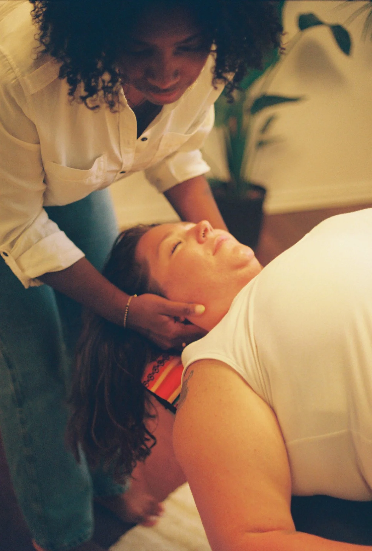 A woman  lying on her back with eyes closed, while a chiropractor with curly hair gives an adjustment in a cozy indoor setting.