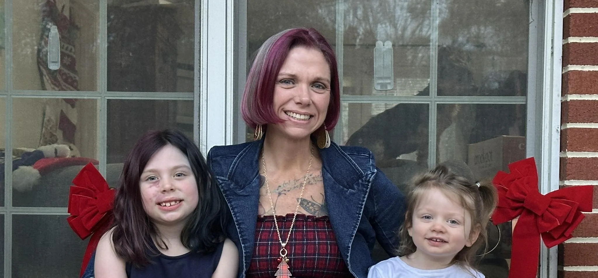 A woman with red hair and two young girls standing outside in front of a window decorated with Christmas bows.
