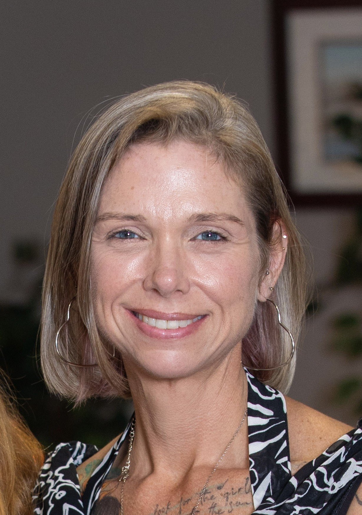 A smiling woman with short blonde hair, blue eyes, wearing hoop earrings and a patterned black and white top, standing indoors.