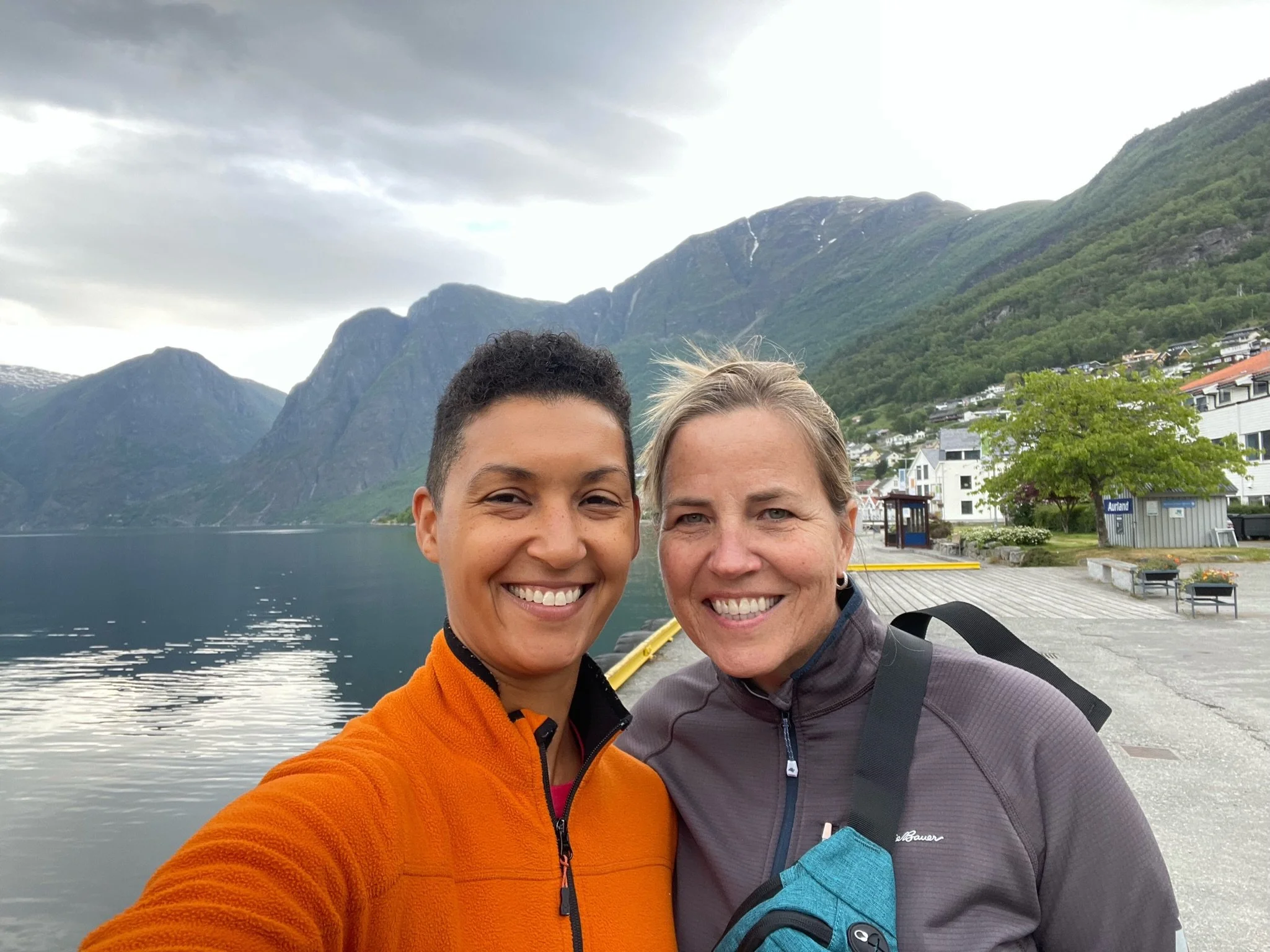 Two women smiling for a selfie in front of a lake surrounded by mountains, with cloudy sky overhead.