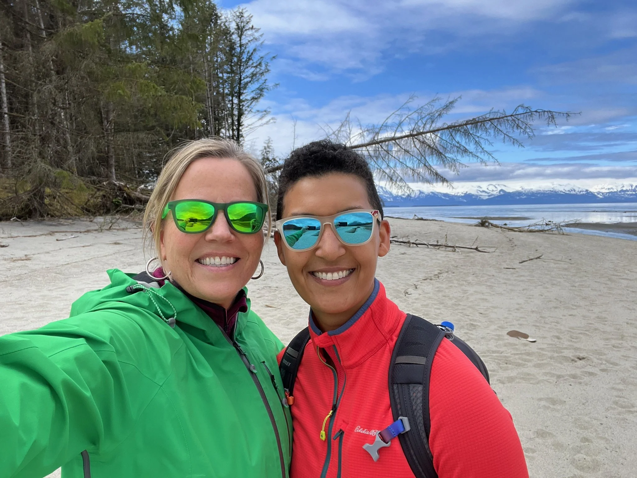 Two women wearing sunglasses and colorful outdoor jackets smiling on a beach with trees, sand, and mountains in the background.