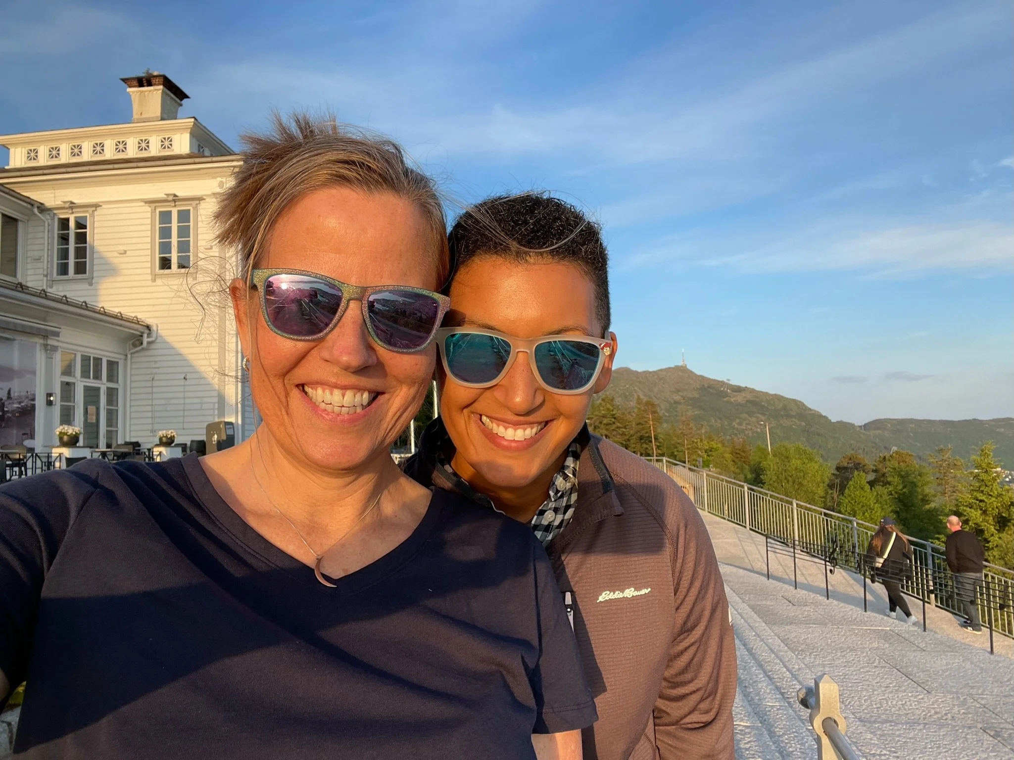 Two smiling women wearing sunglasses taking a selfie outdoors with a mountain and blue sky in the background.