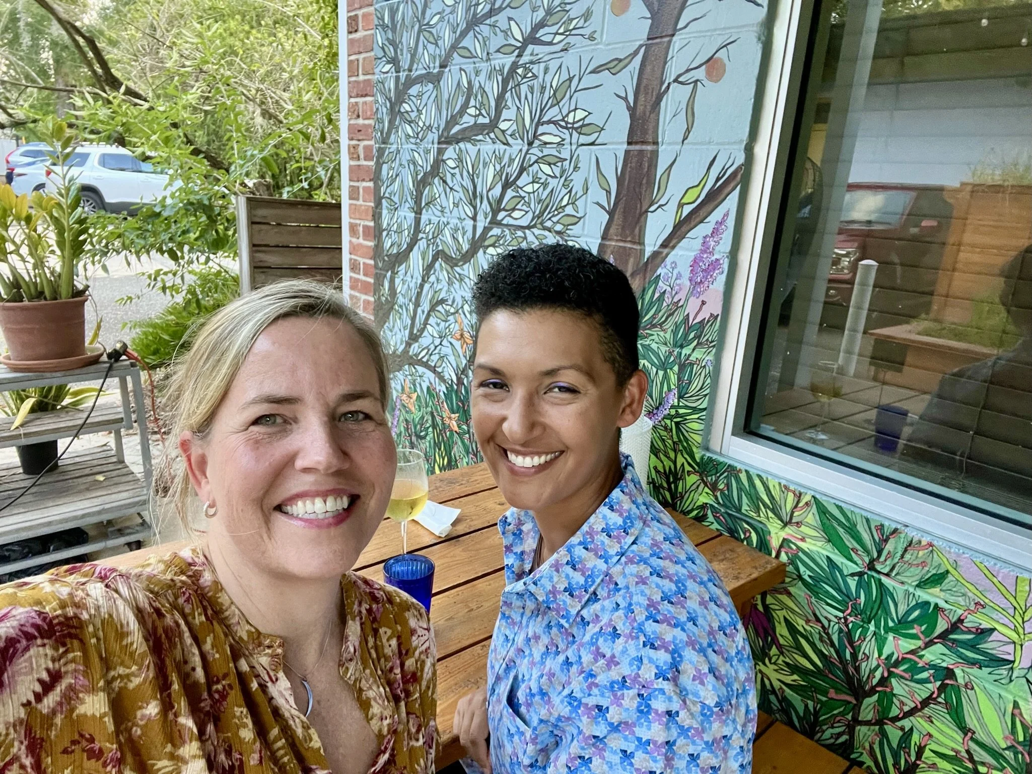 Two women taking a selfie together at an outdoor table. One has blonde hair and the other has short black hair. They are smiling, and there are drinks on the table. Behind them is a mural of a tree on a brick wall.