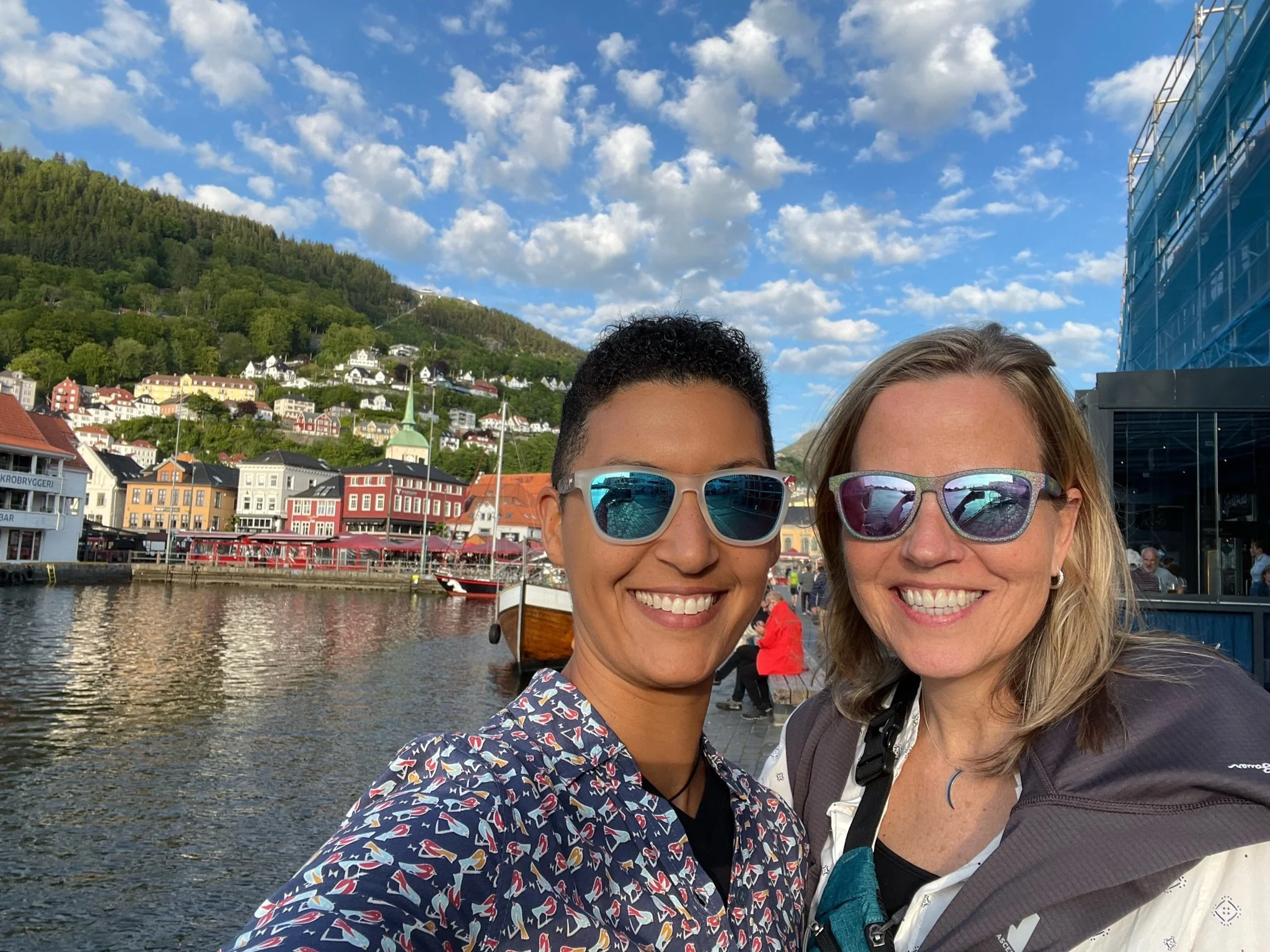 Two women wearing sunglasses taking a selfie together outdoors near a waterfront with colorful buildings, a church with a tall steeple, and green hills under a partly cloudy sky.