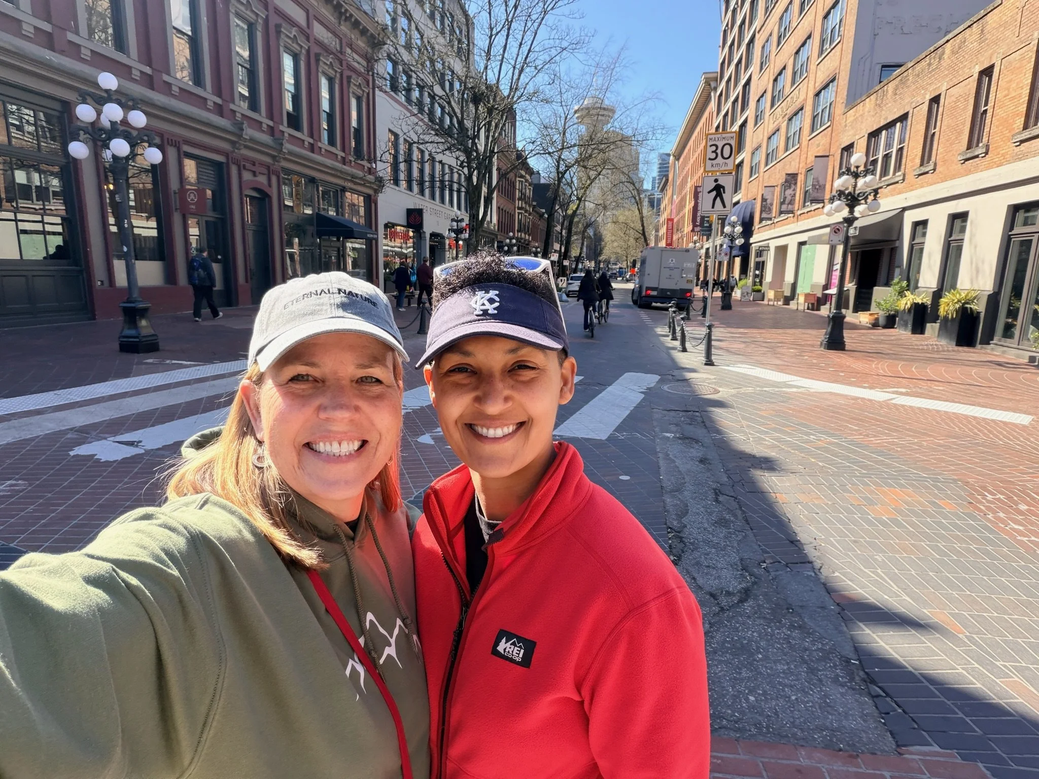 Two smiling women taking a selfie on a city street with historic buildings, street lamps, pedestrians, and cyclists in the background on a sunny day.