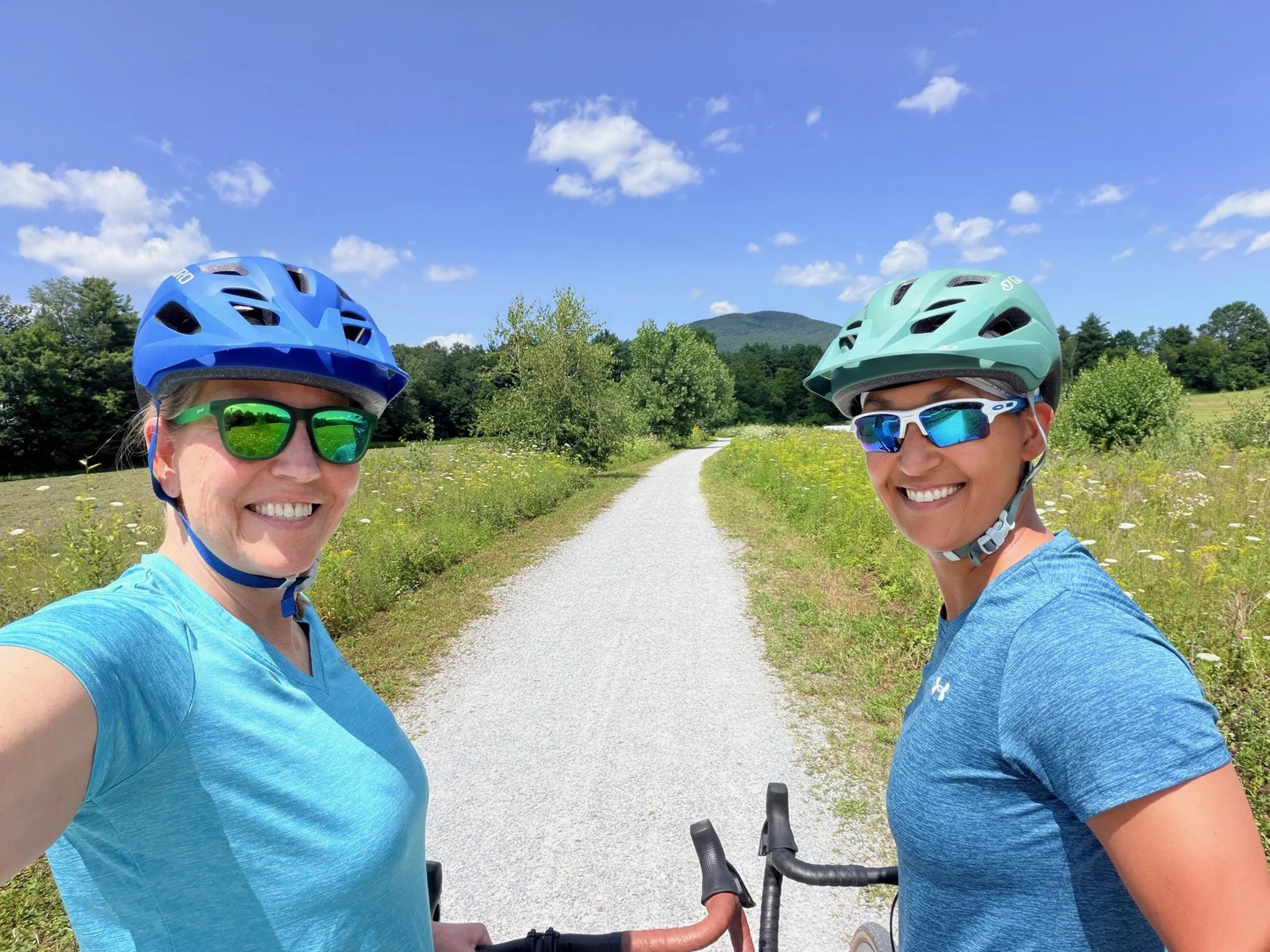 Two women wearing biking helmets and sunglasses, smiling on a sunny day in a natural outdoor setting with a dirt path, green trees, bushes, and a mountain in the background.