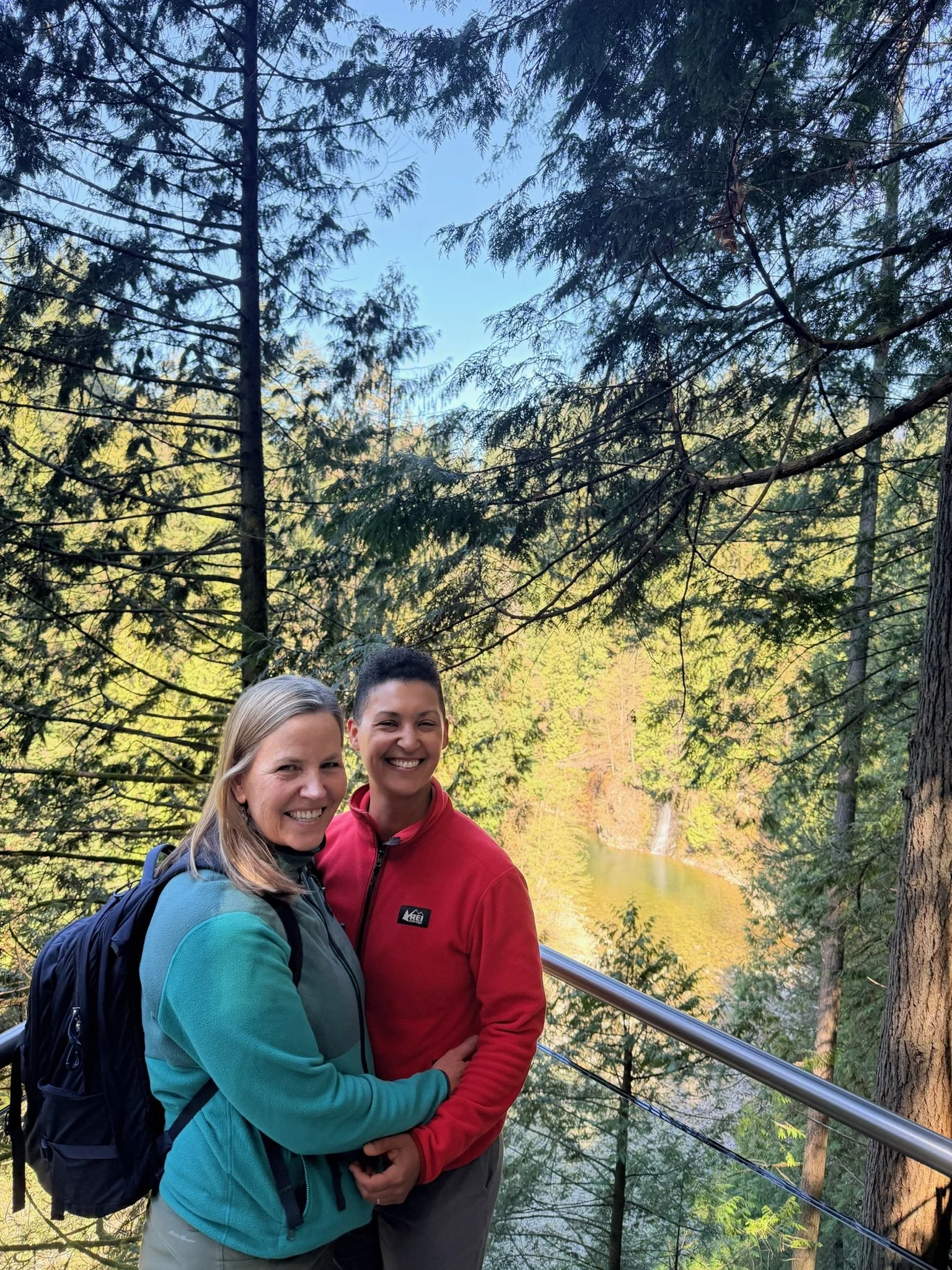 Smiling women and man standing outdoors among tall trees, wearing hiking clothing and backpacks, near a lake with a lush forest in the background.