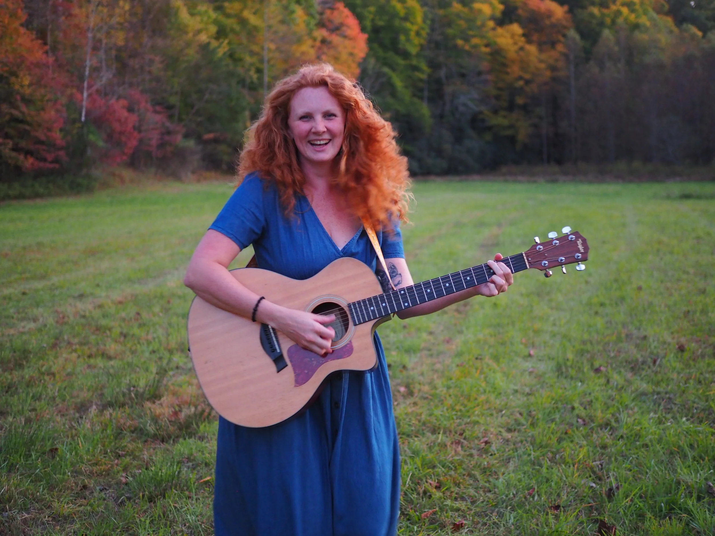 A woman with curly red hair wearing a blue dress playing an acoustic guitar outdoors during autumn, with colorful trees in the background.