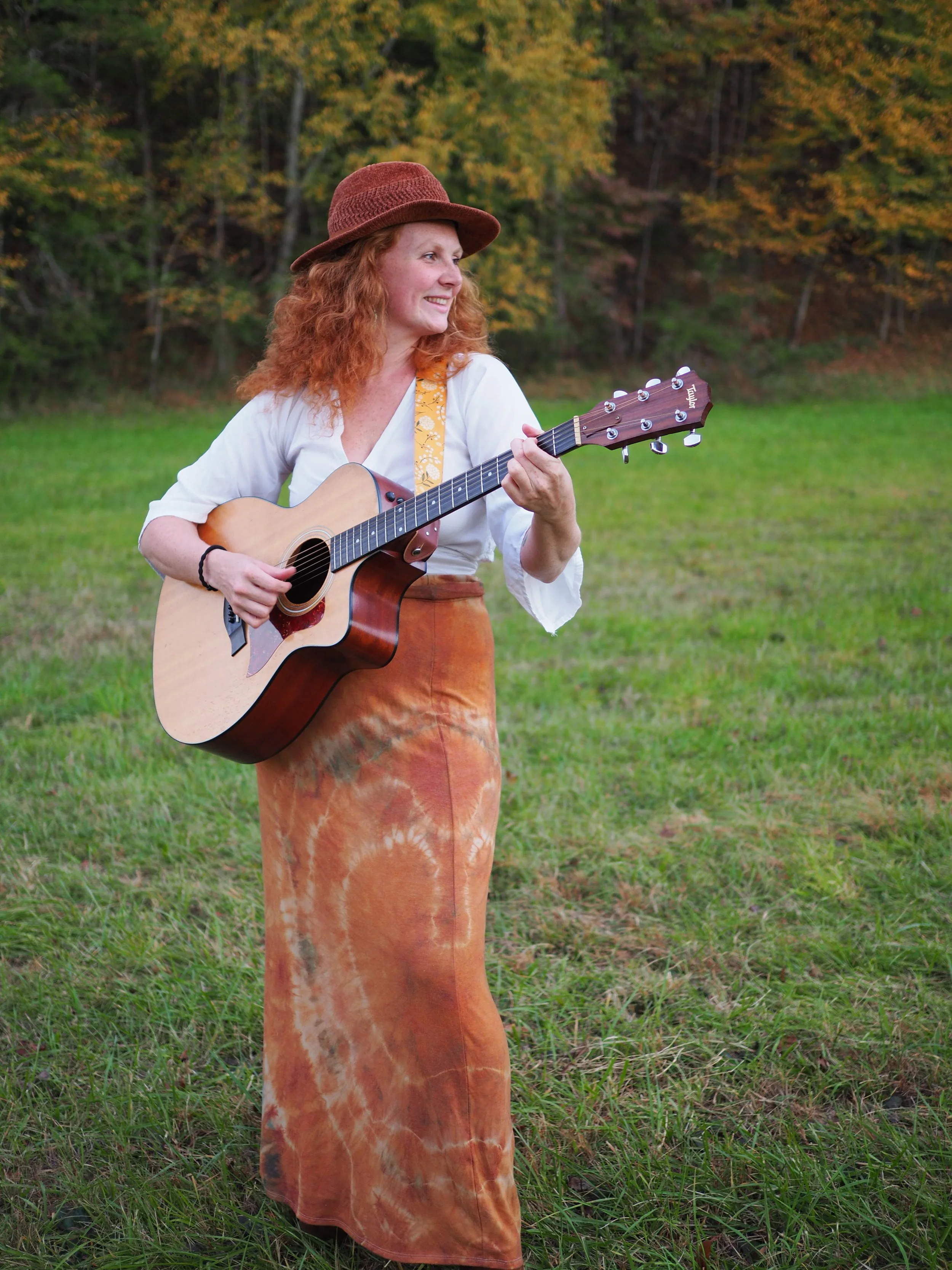 A woman with curly red hair playing an acoustic guitar outdoors in a grassy field with trees displaying fall foliage in the background.