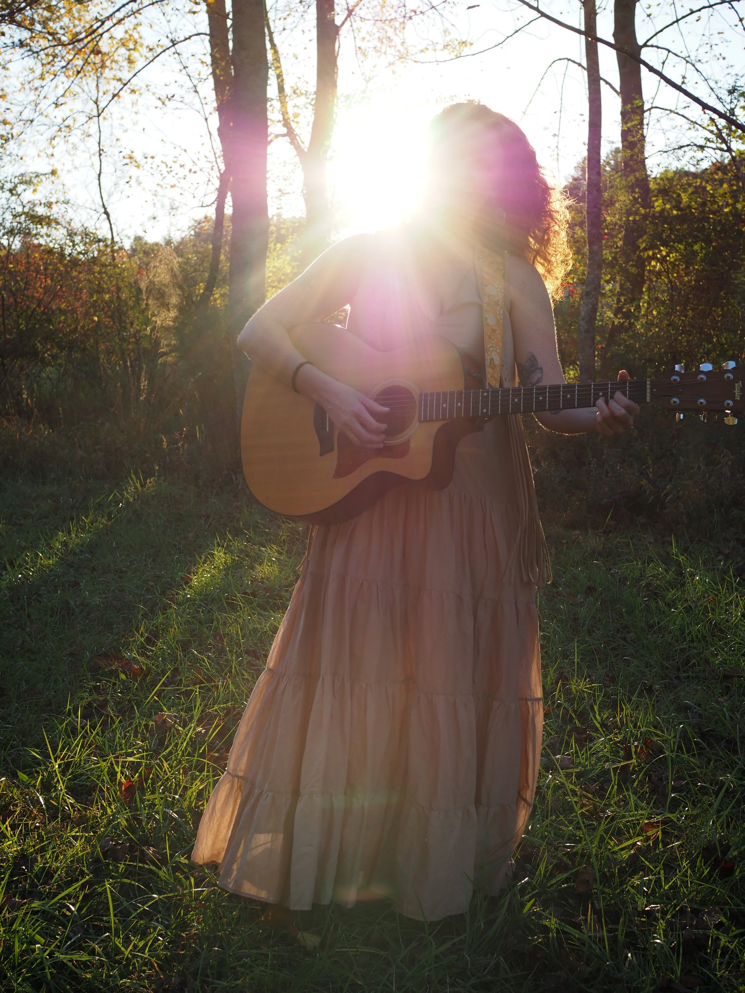 A woman with curly hair wearing a long, beige dress and earrings is playing an acoustic guitar outdoors in a wooded area during sunset, with sunlight shining behind her.