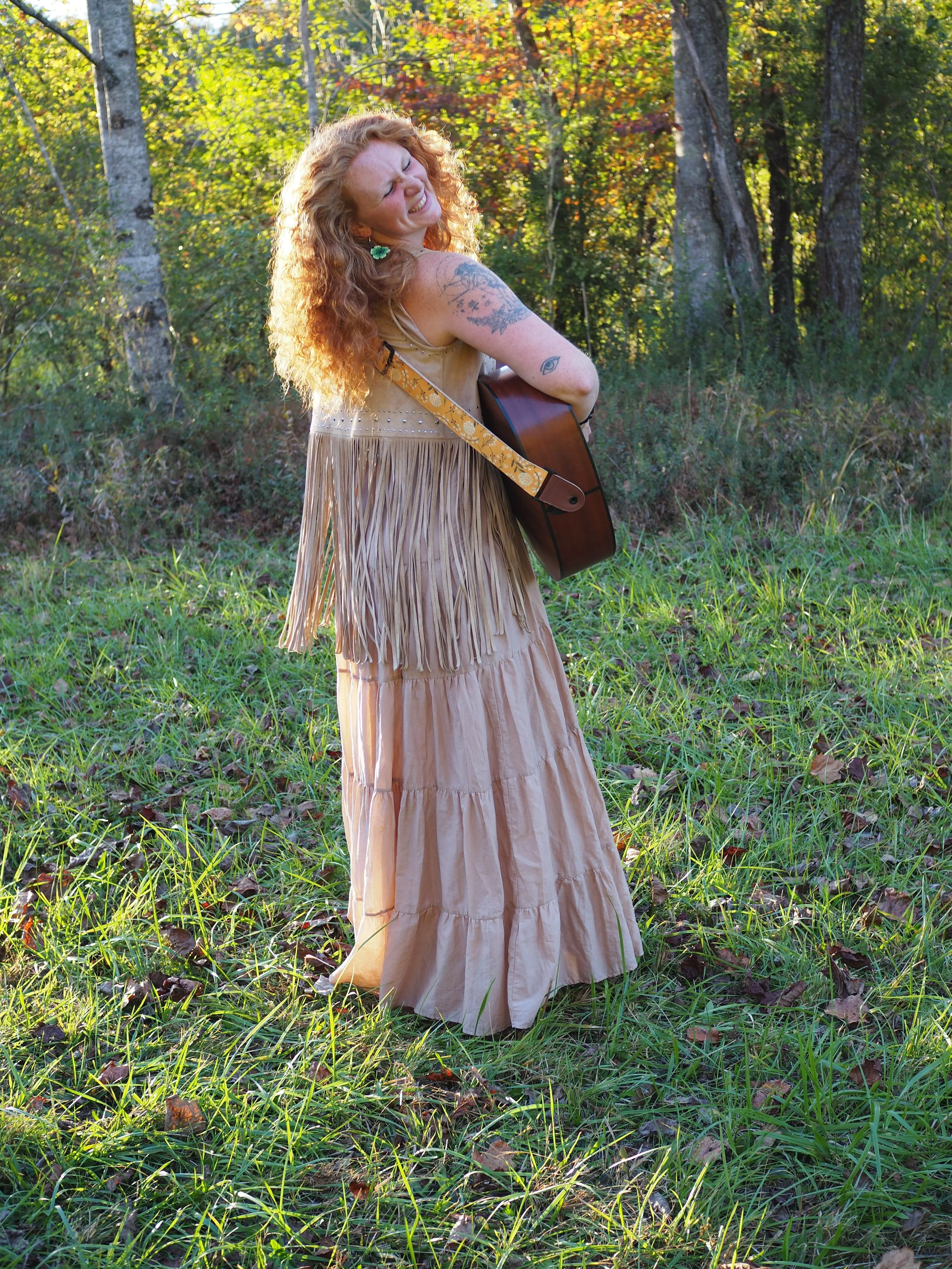 A woman with curly red hair smiling and playing guitar in a forest clearing with green grass and autumn-colored leaves on trees.