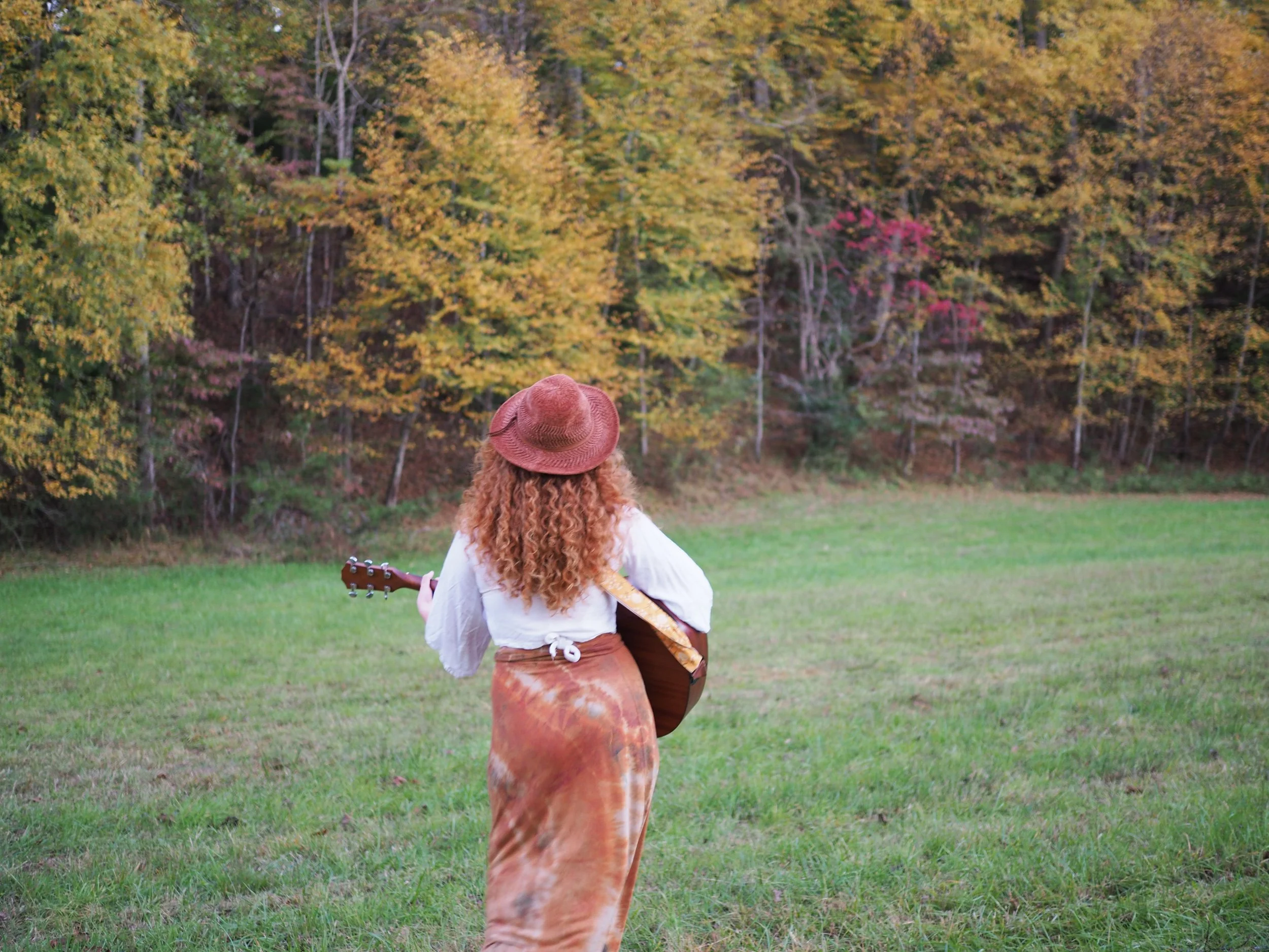 A woman with curly red hair, wearing a brown wide-brim hat, white shirt, and patterned pants, stands in a grassy field playing an acoustic guitar, with colorful autumn trees in the background.