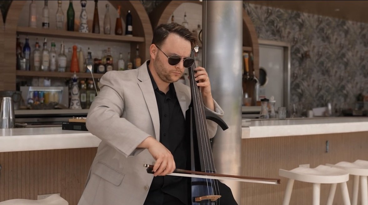 Man wearing sunglasses and a beige blazer playing a cello at a bar or restaurant counter.
