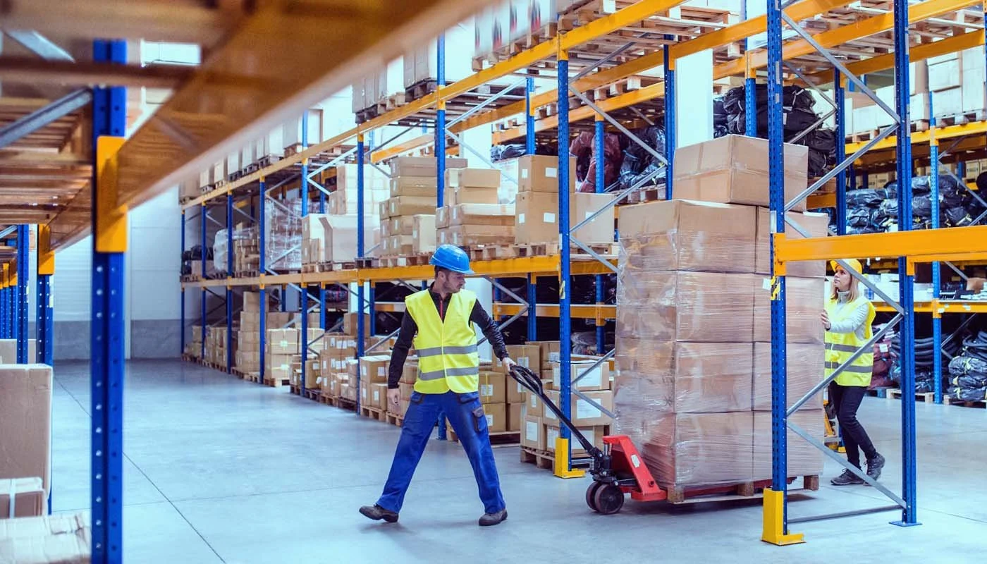 Warehouse workers in yellow safety vests and blue helmets moving pallets of boxes with a pallet jack in a large storage facility with metal shelves filled with boxes and packages.