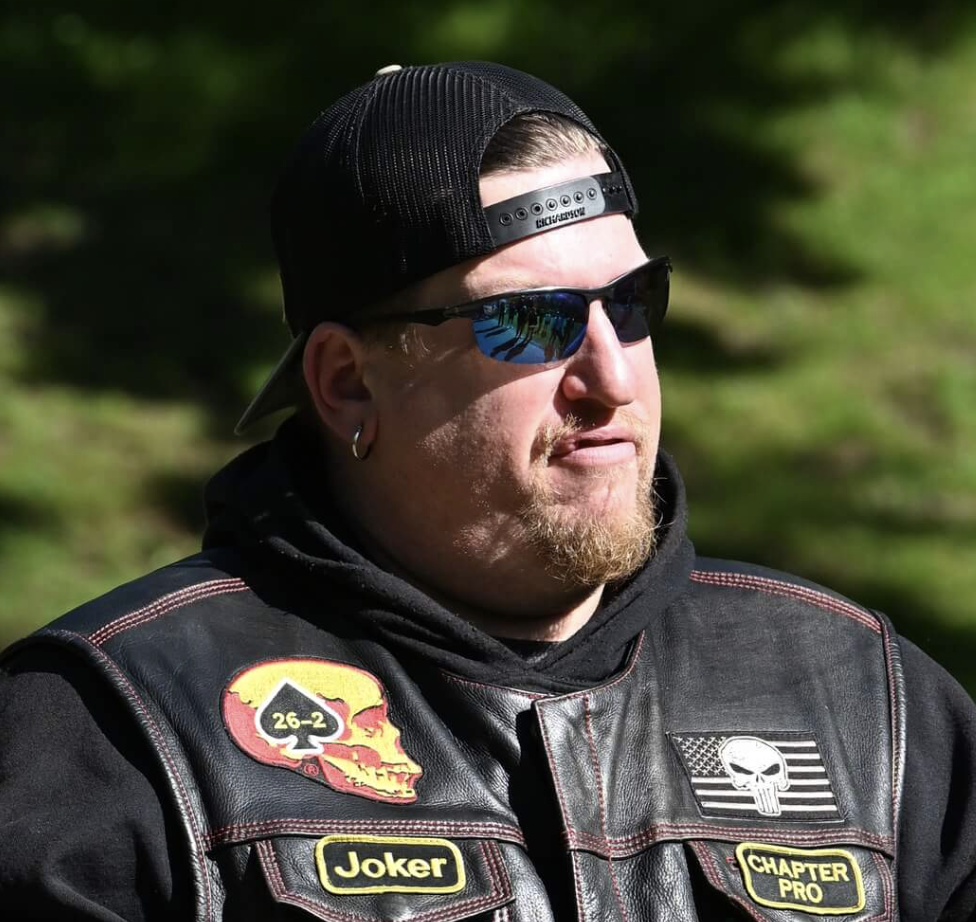A man wearing a black cap backward, tinted sunglasses, earrings, and a leather vest with patches, outdoors with a green background.