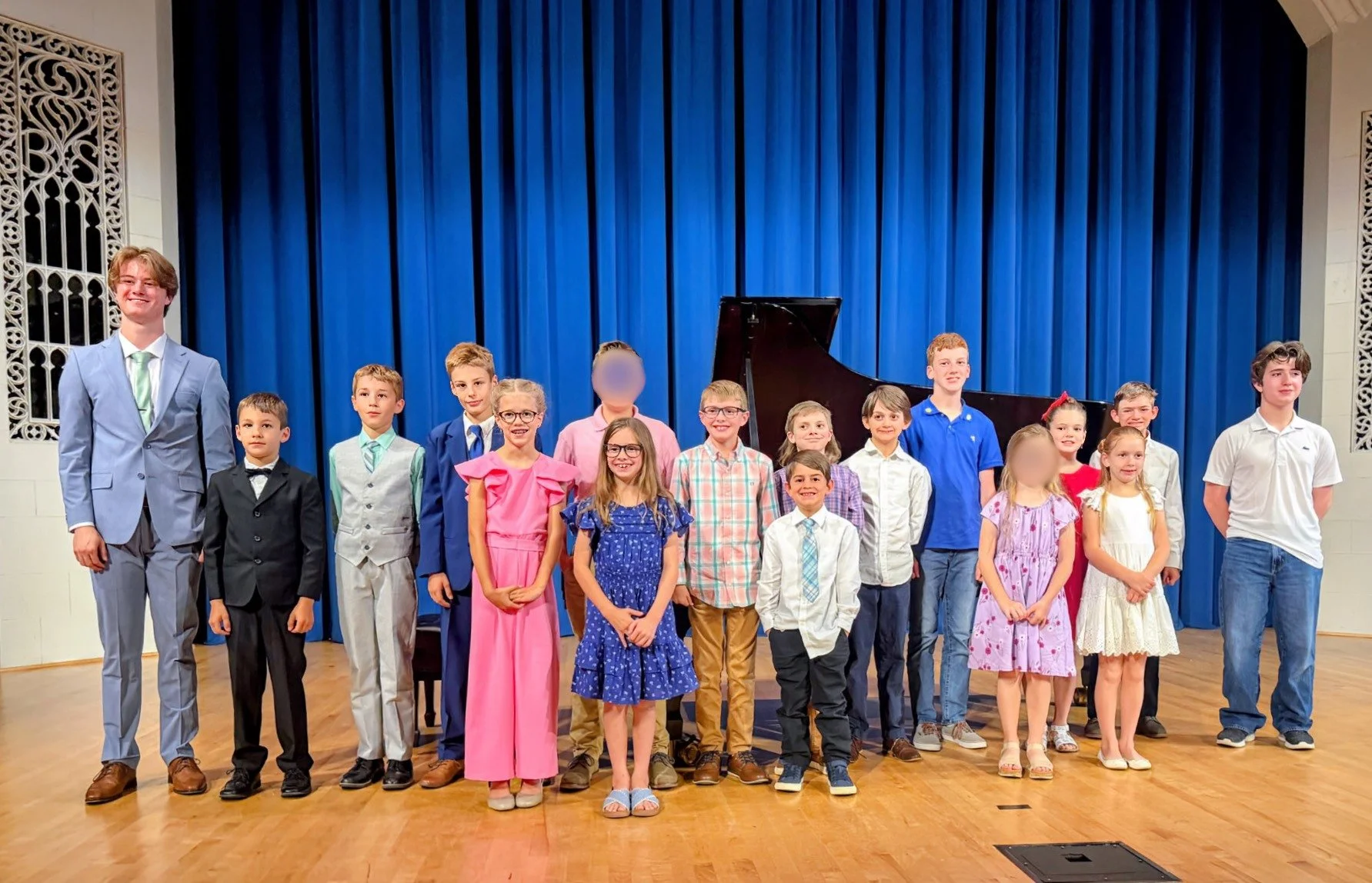 A group of children and their teacher, standing on stage in front of a grand piano and a blue curtain. The children are dressed in formal and colorful attire and are smiling.