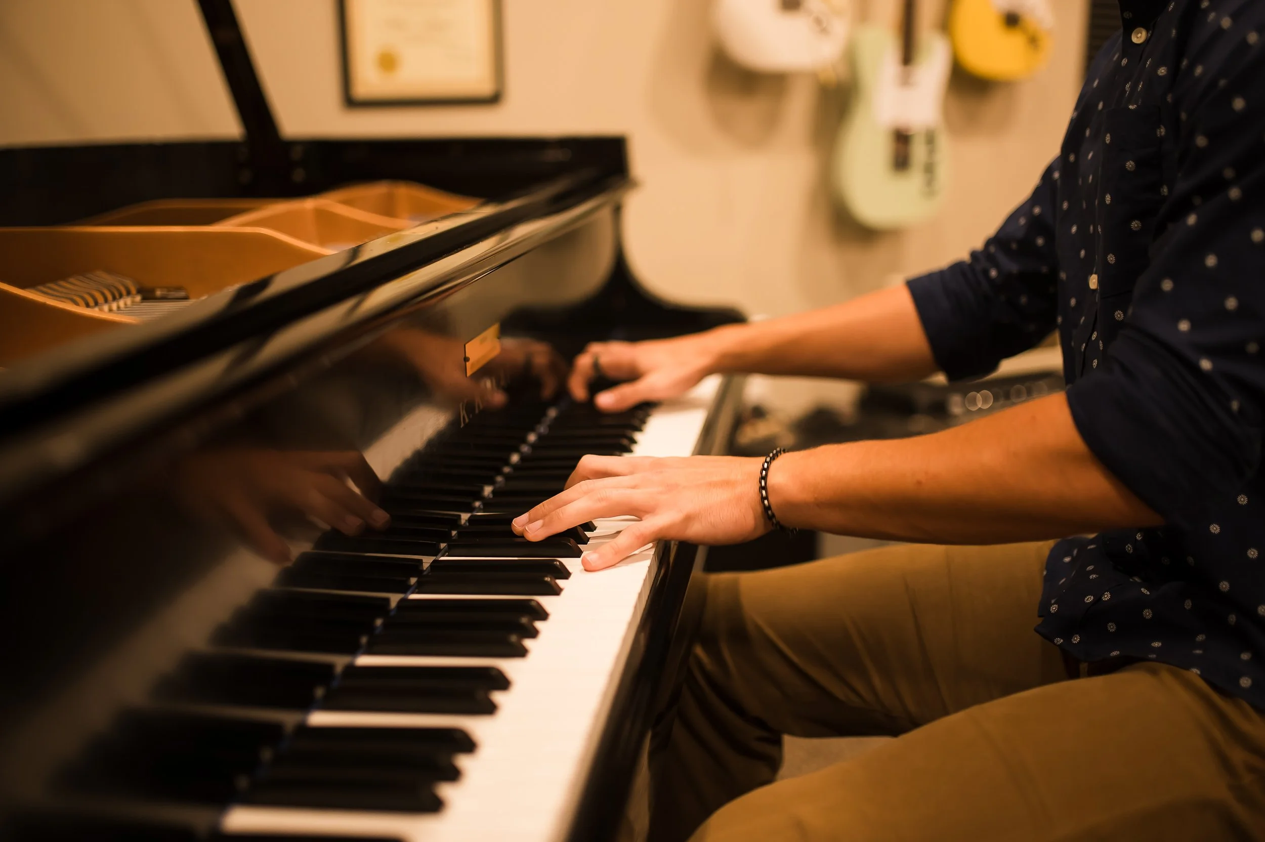 A person playing a grand piano indoors, with a certificate and guitars hanging on the wall in the background.